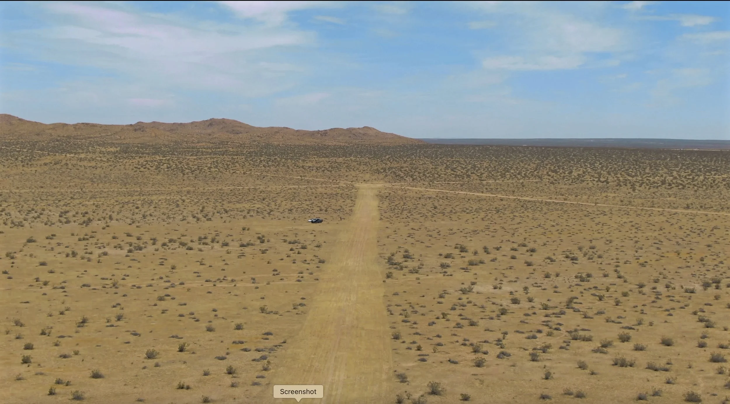 A vast desert landscape with sparse bushes, a dirt runway stretching into the distance, low mountains on the horizon, and a blue sky with some clouds.