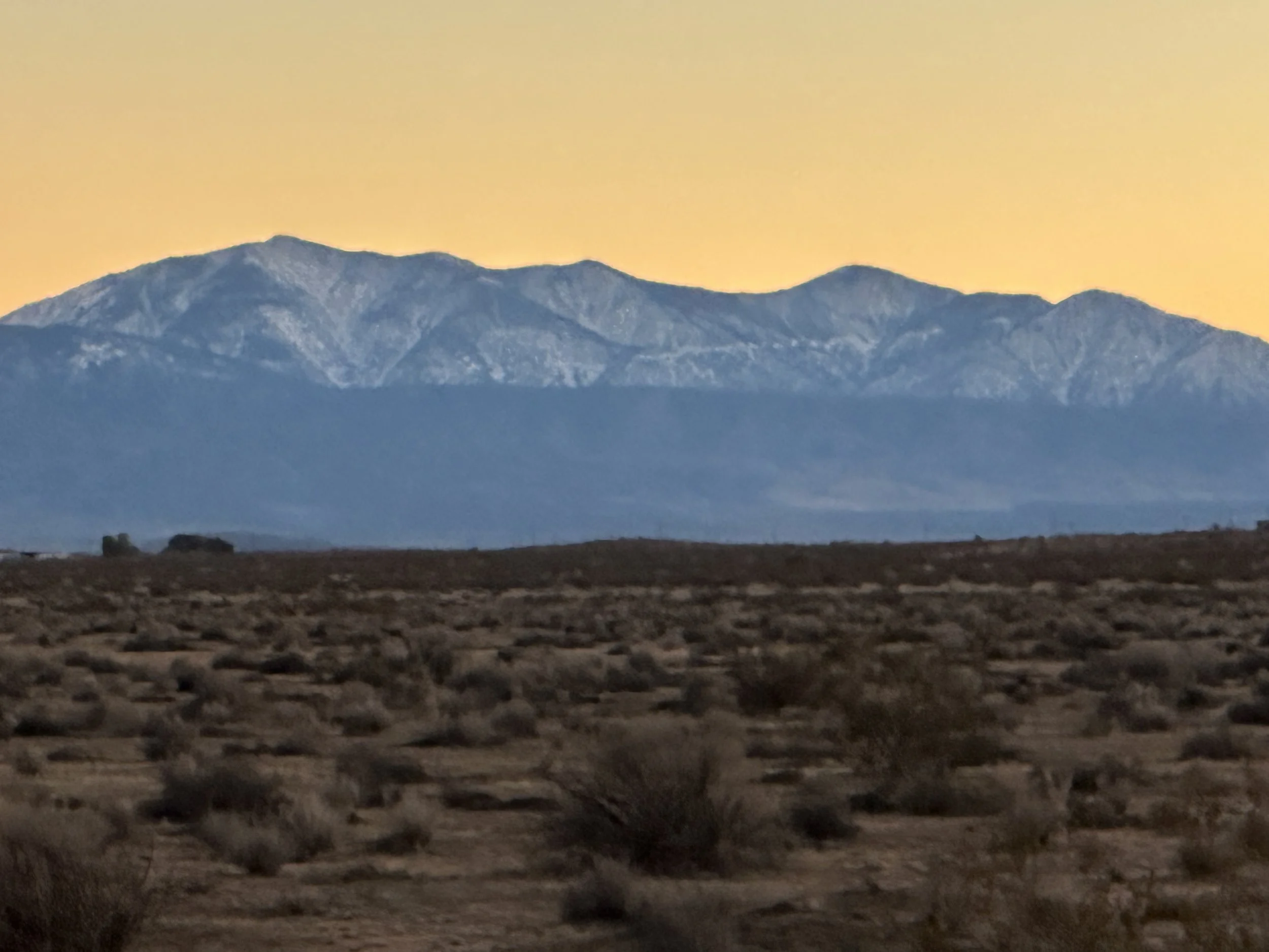 A mountain range with snow-capped peaks against a yellowish sunset sky, foreground shows a barren desert landscape with scattered bushes.
