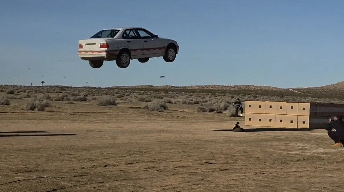 A car appears to be floating in mid-air in a desert landscape while people and large boxes are nearby.