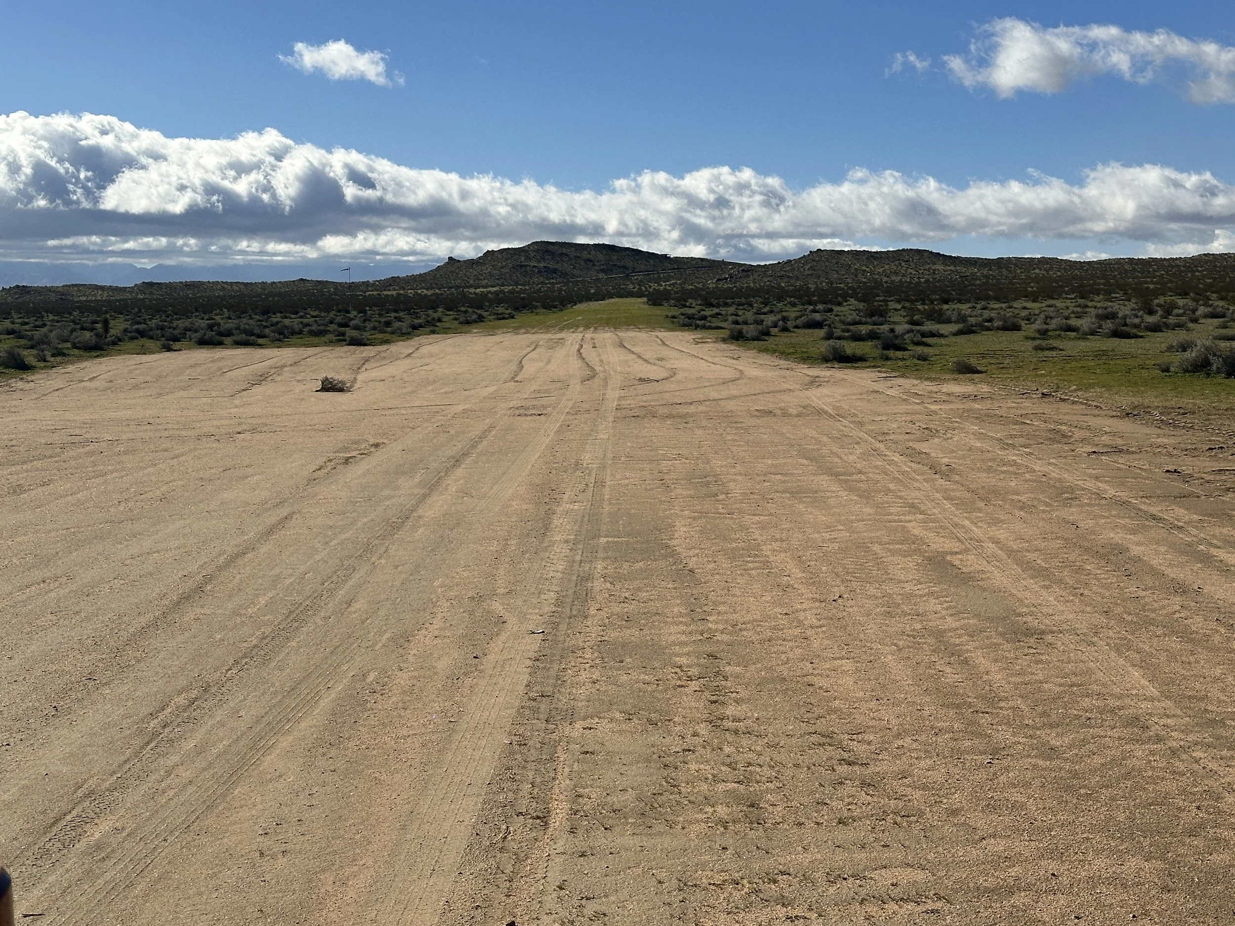 Dirt road extending into the distance with hills and desert shrubs under a partly cloudy sky.