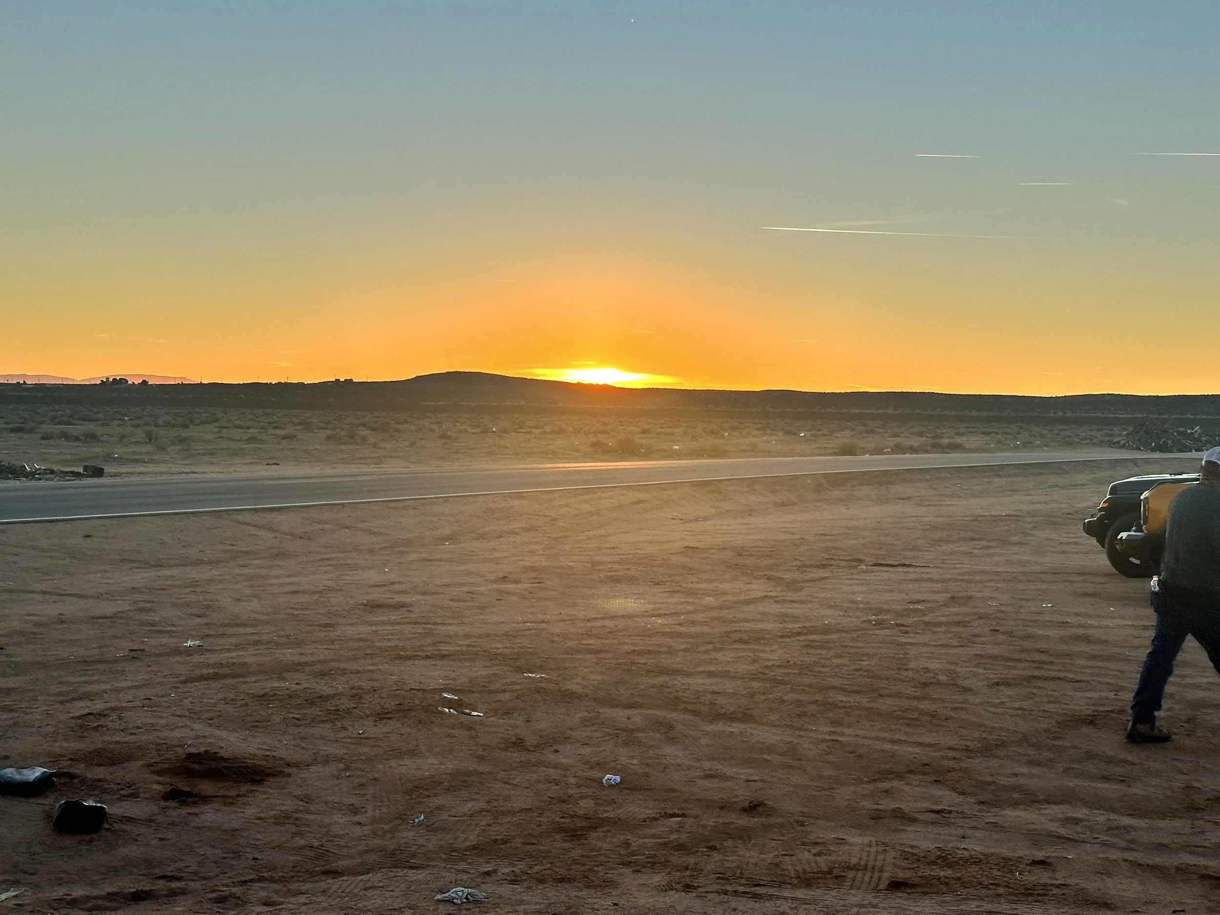 A sunset over a desert landscape with a clear sky, a road, and parked cars on the right side. A person is standing near the cars, looking toward the sunset.
