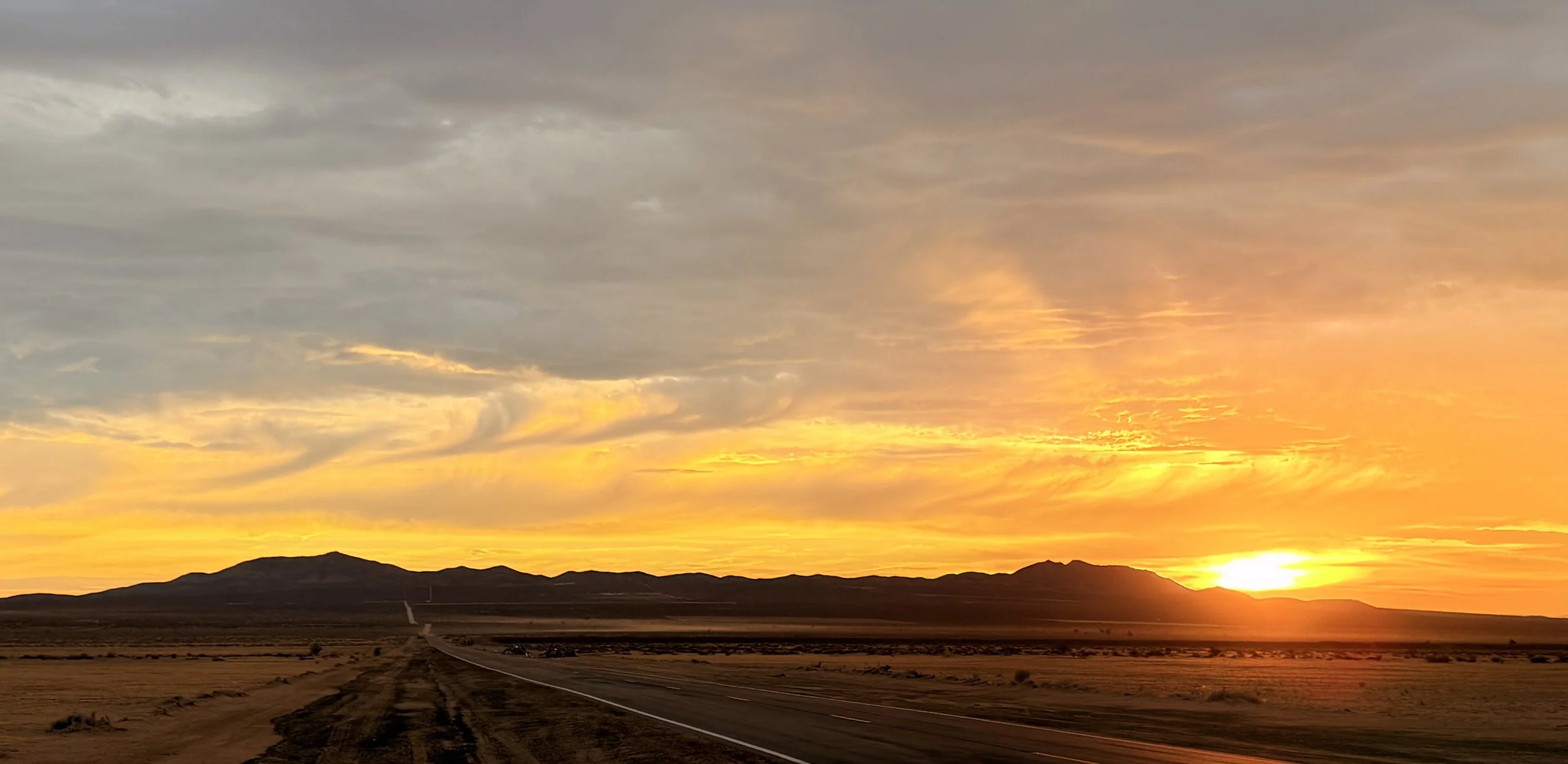A sunset over a desert landscape with a dirt road leading towards distant mountains and a mix of clouds and vibrant orange and yellow colors in the sky.