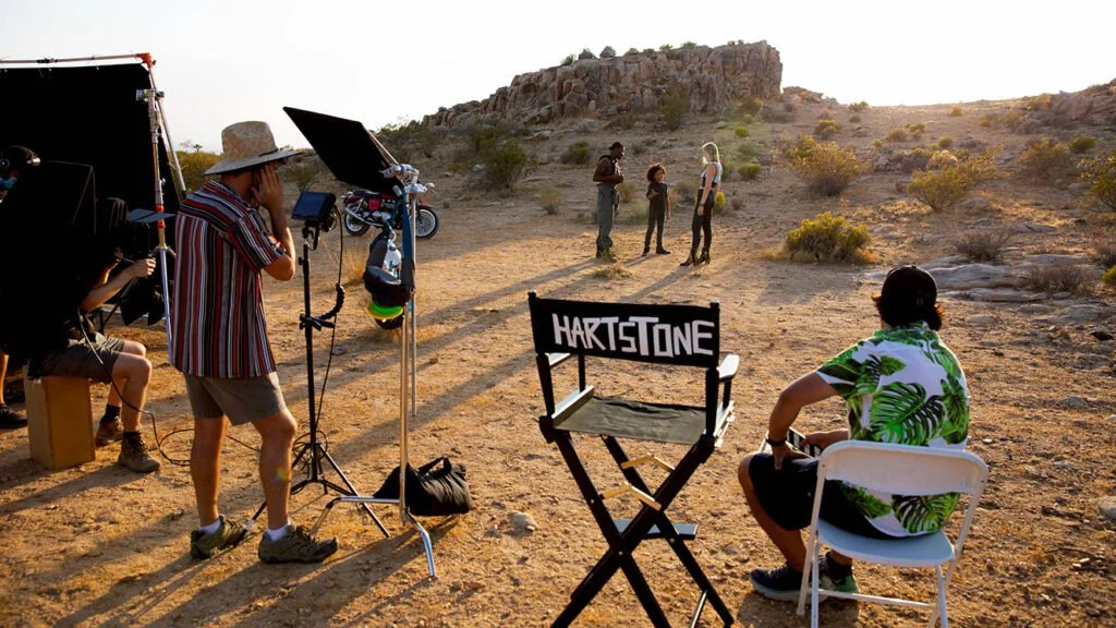 A behind-the-scenes view of a film or photo shoot in a desert landscape at sunset, with crew members, a director's chair labeled 'HARTSTONE', and actors in the background.