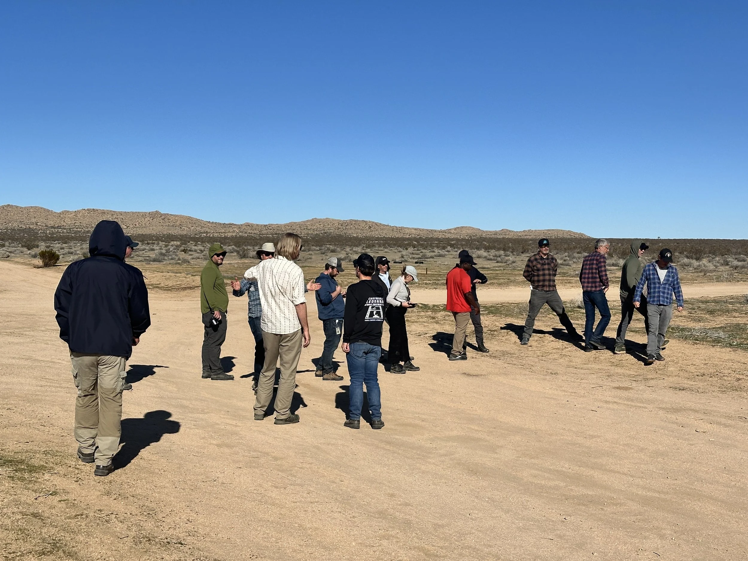 Group of people walking on a dirt path in a desert landscape with mountains in the distance and a clear blue sky.