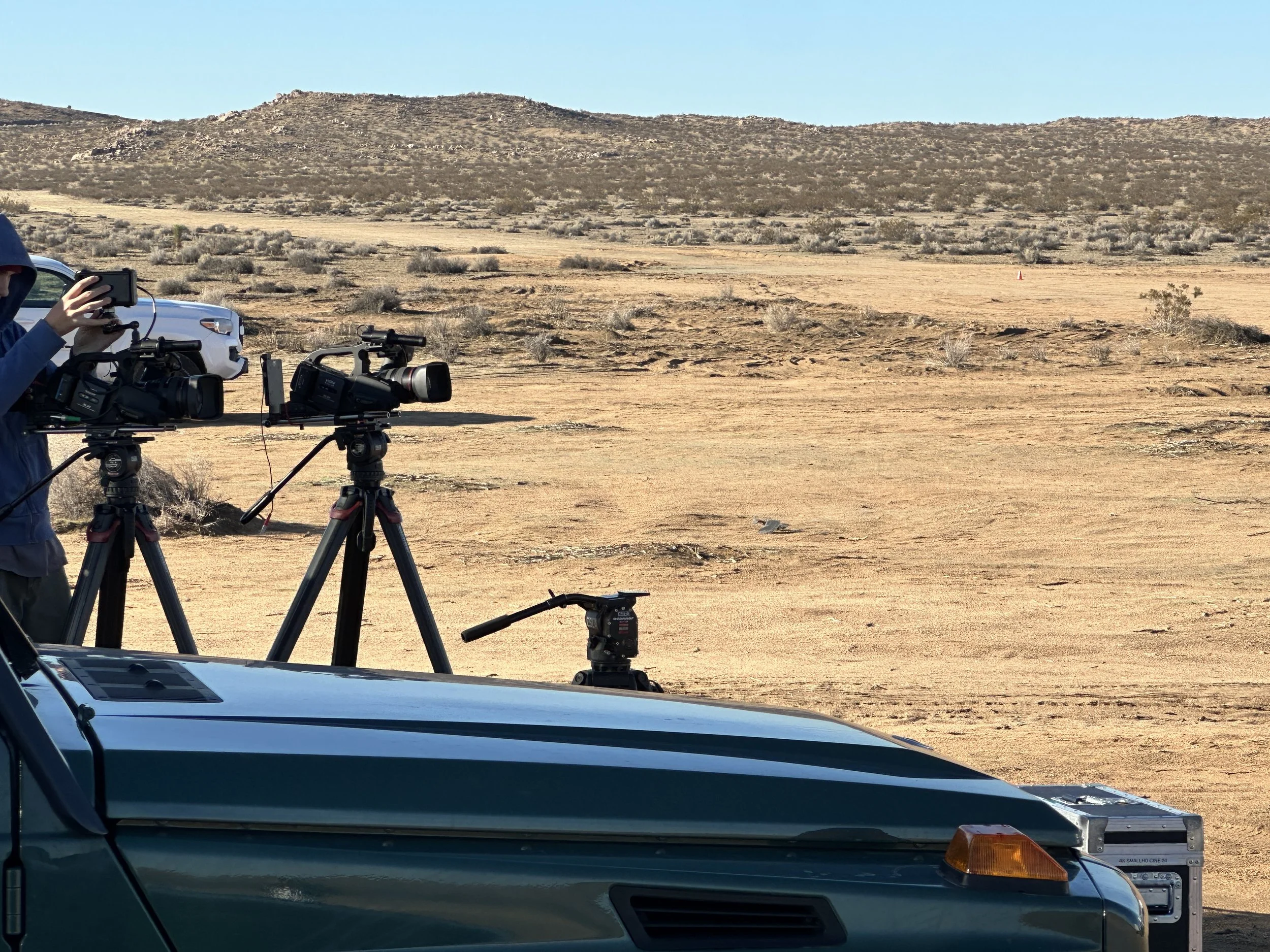 Camera operators filming in a desert landscape with sparse vegetation and distant hills under a clear blue sky.