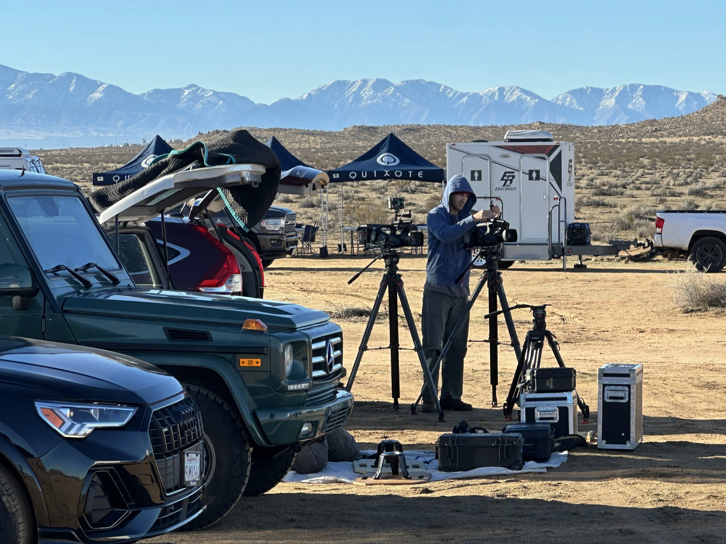 Filmmaker setting up camera equipment in a desert with parked vehicles and mountains in the background.