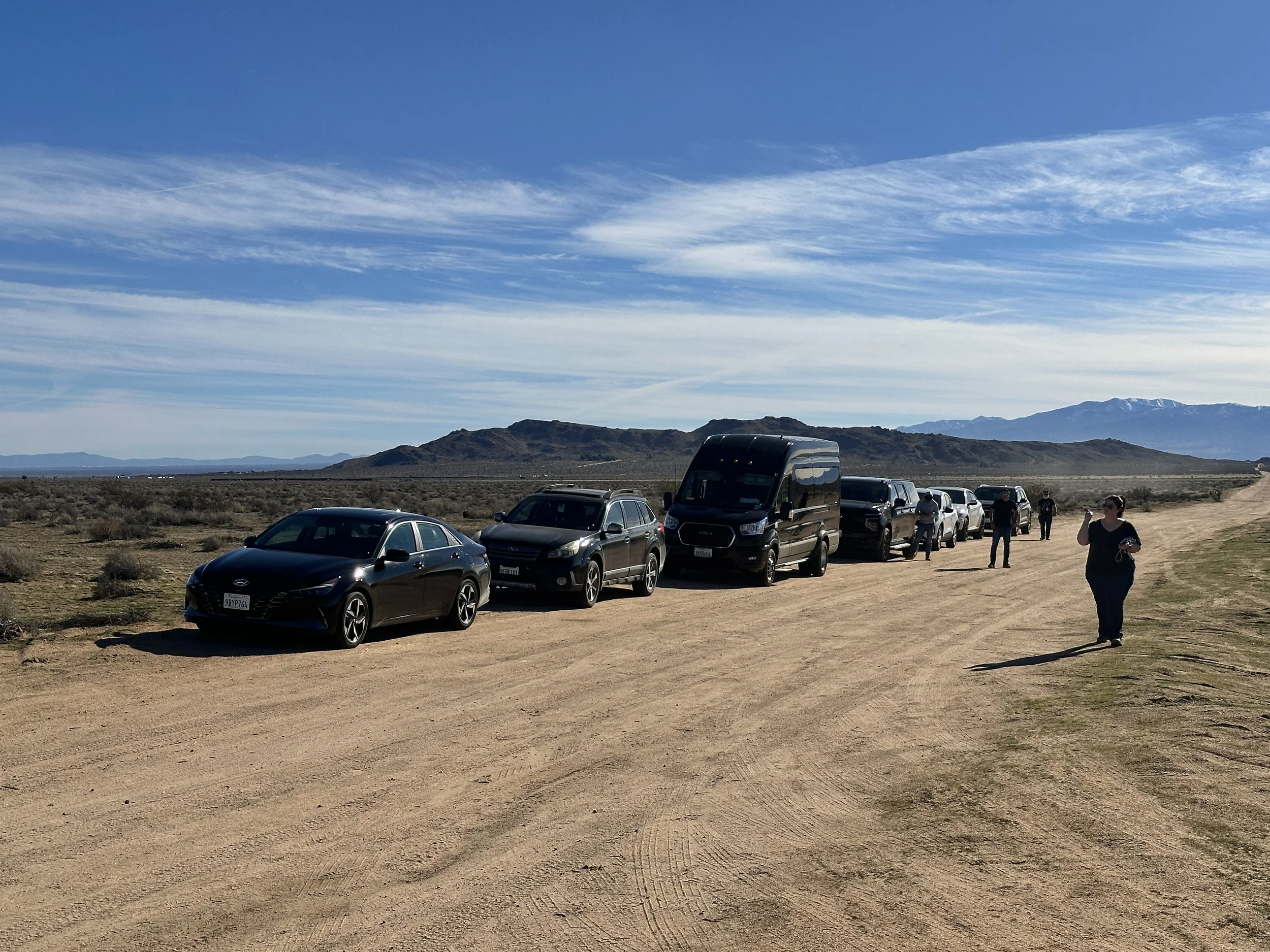 Multiple cars parked along a dirt road in a desert landscape with distant mountains and a partly cloudy sky, and several people walking or standing.