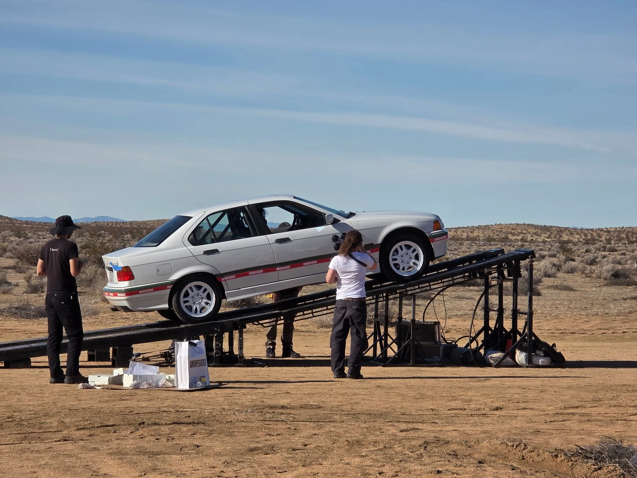 A silver car mounted on a camera rig in a remote desert area with three people around it, one of whom is adjusting something on the car.