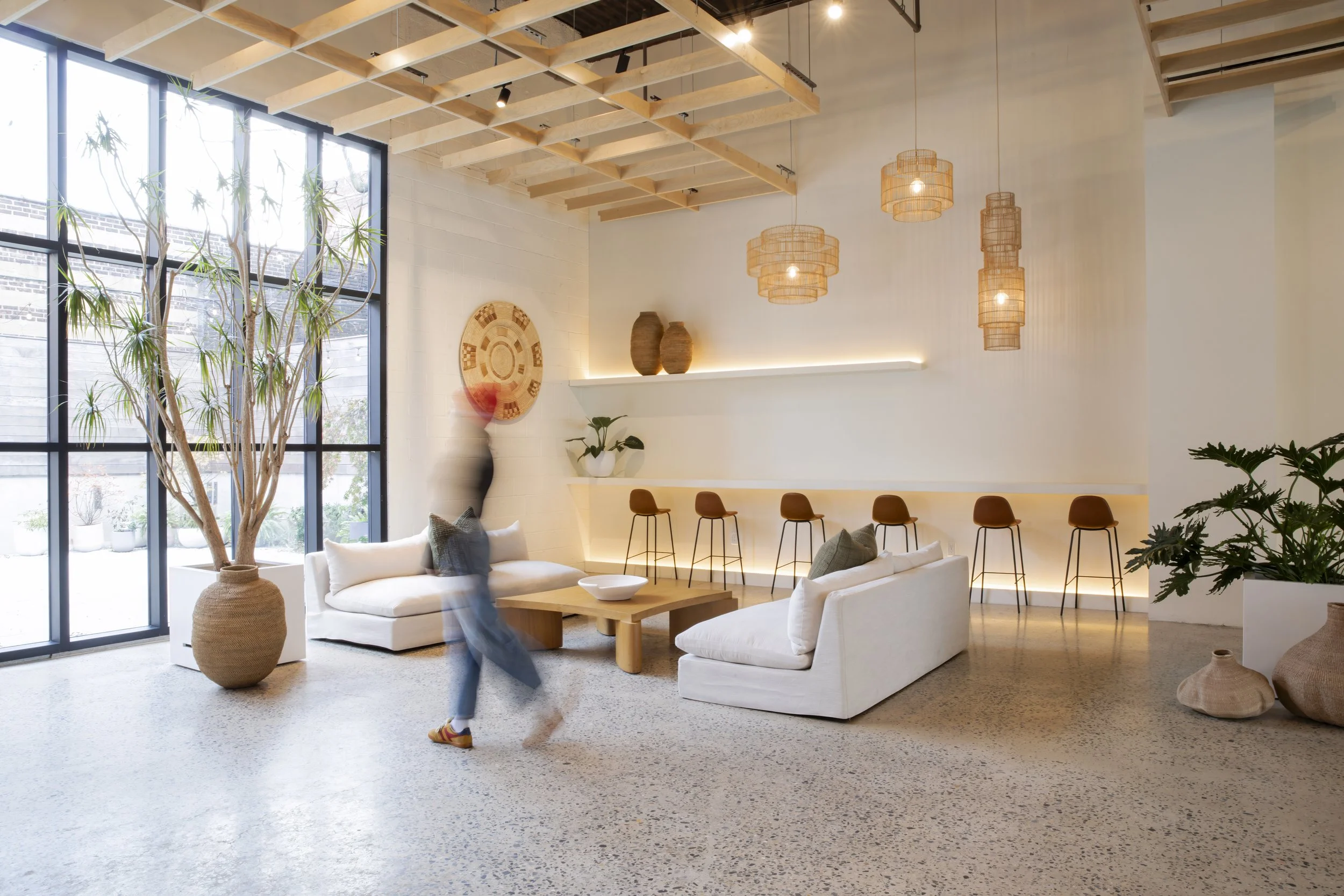 A modern, spacious lounge area with white sofas, a large potted plant, wooden ceiling beams, and hanging wicker lamps. Barstools line the back wall with a shelf and decorative vases. Large windows let in natural light.