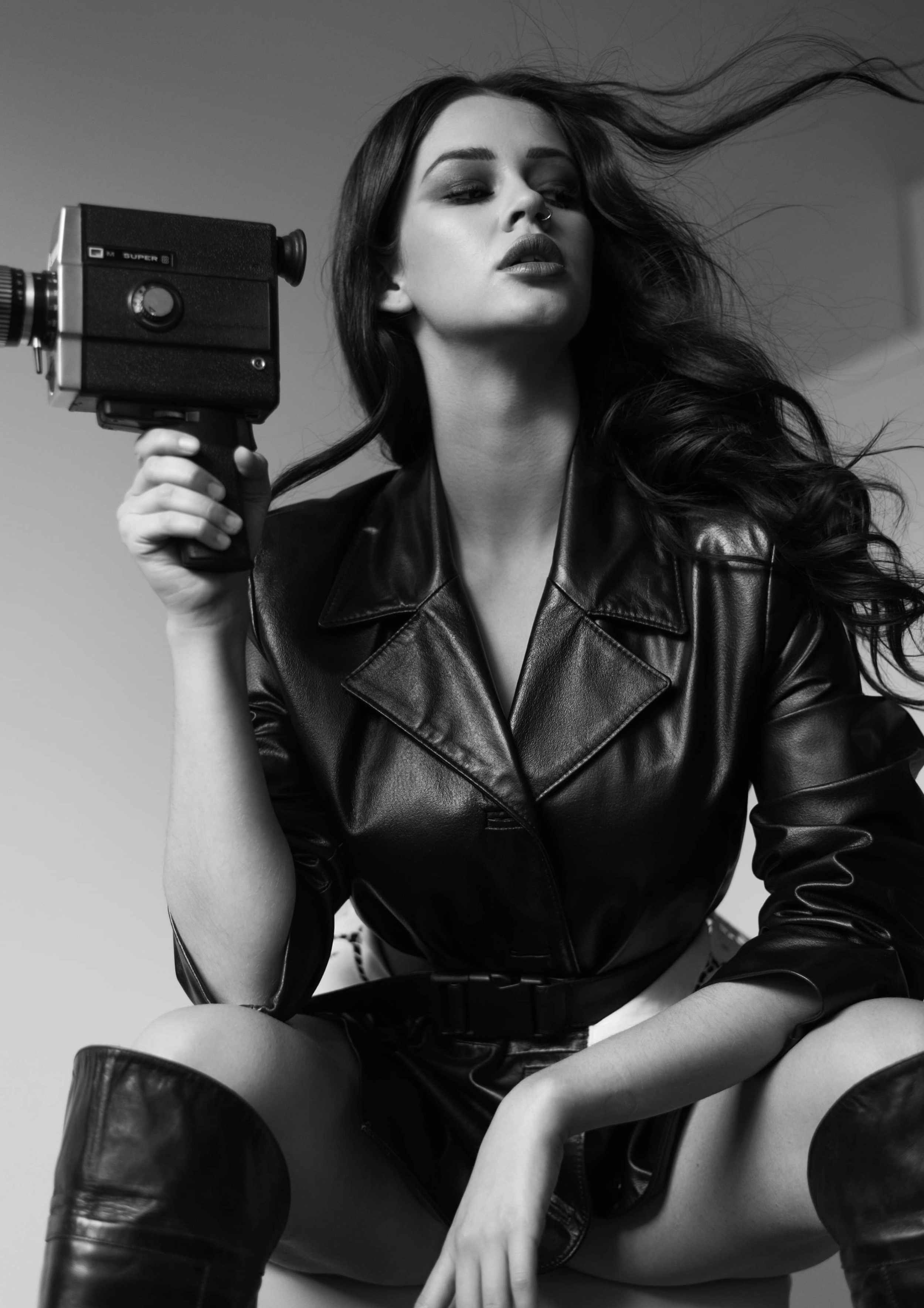 Black and white photo of a woman with long hair, wearing a leather jacket and boots, holding a vintage film camera, with her hair blowing.