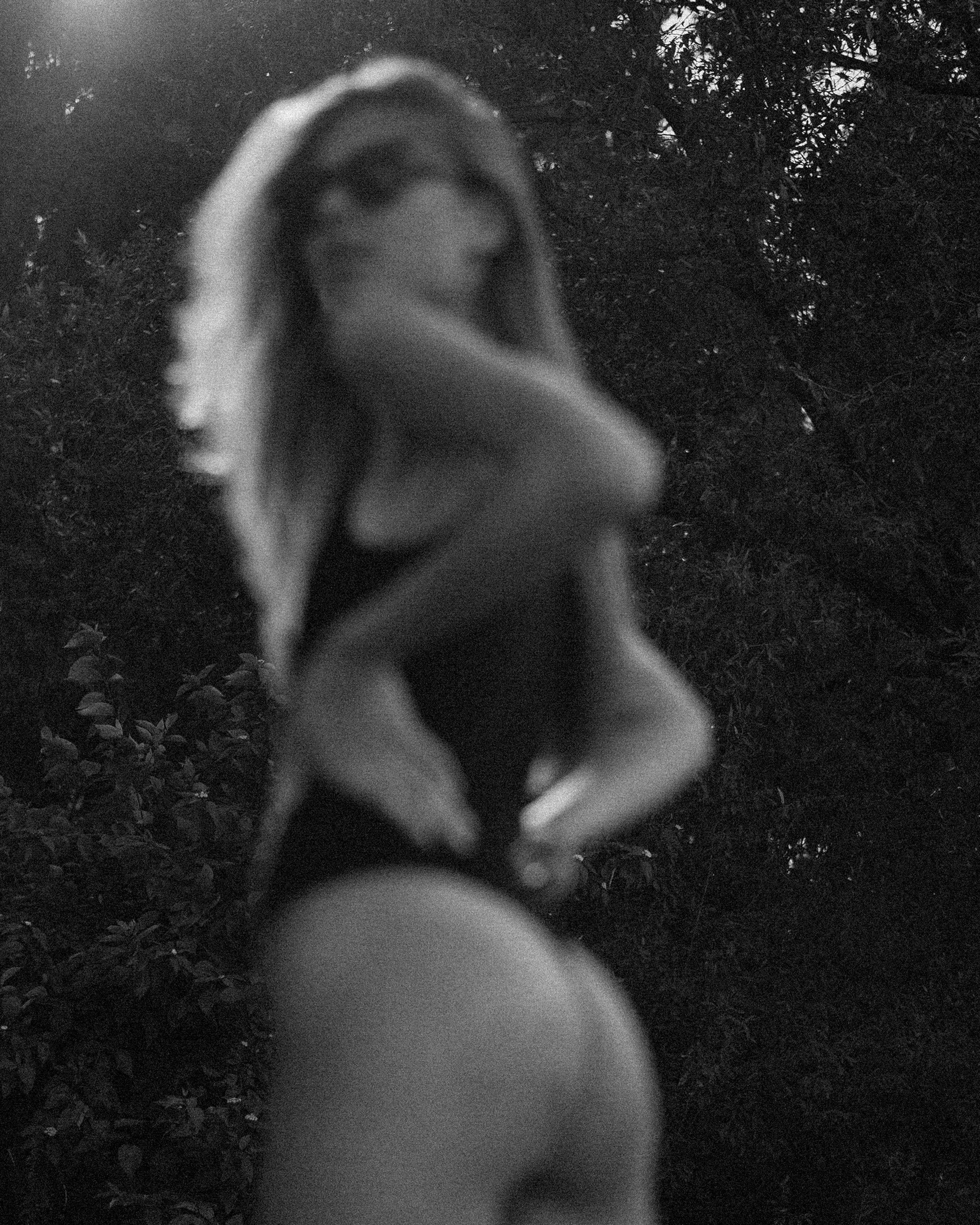 Blurry black and white photo of a woman outdoors, wearing a swimsuit and a towel draped over her shoulder, with trees in the background.