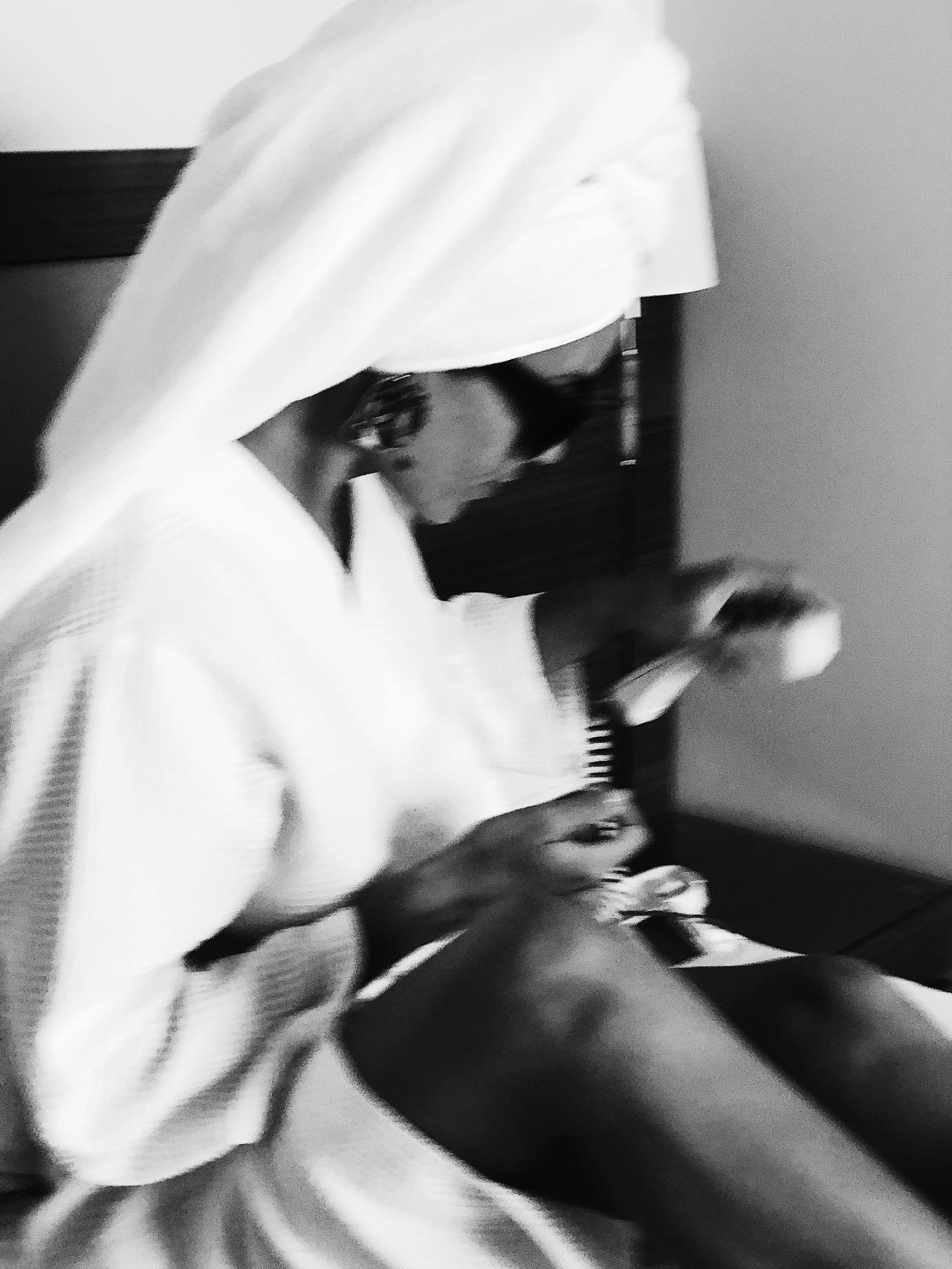 A woman wrapped in a towel with a towel on her head, sitting on a bed, holding a phone, with another woman sitting nearby in a well-lit room.