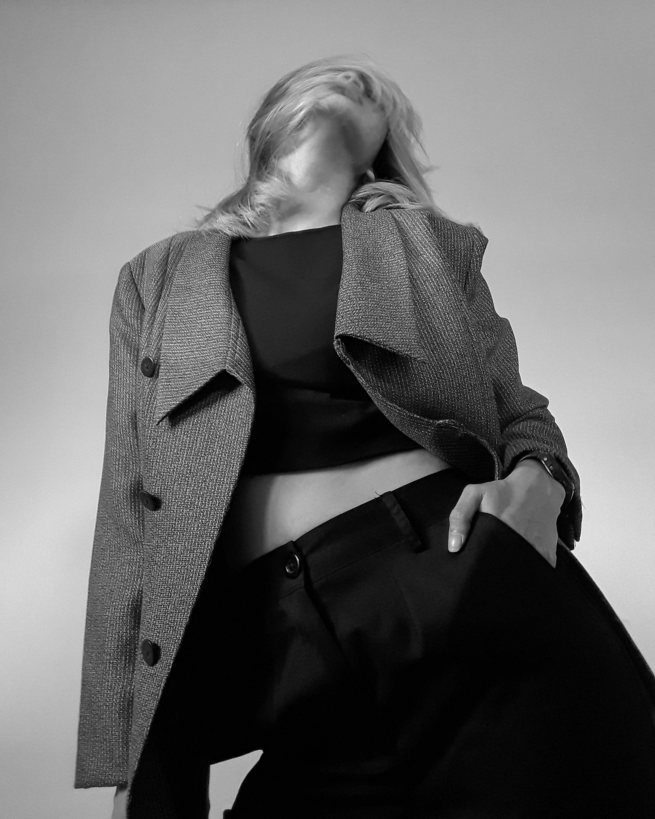 Low-angle black and white photo of a woman in a blazer, crop top, and pants, posing with her hand in her pocket, head tilted back and hair partially covering her face.