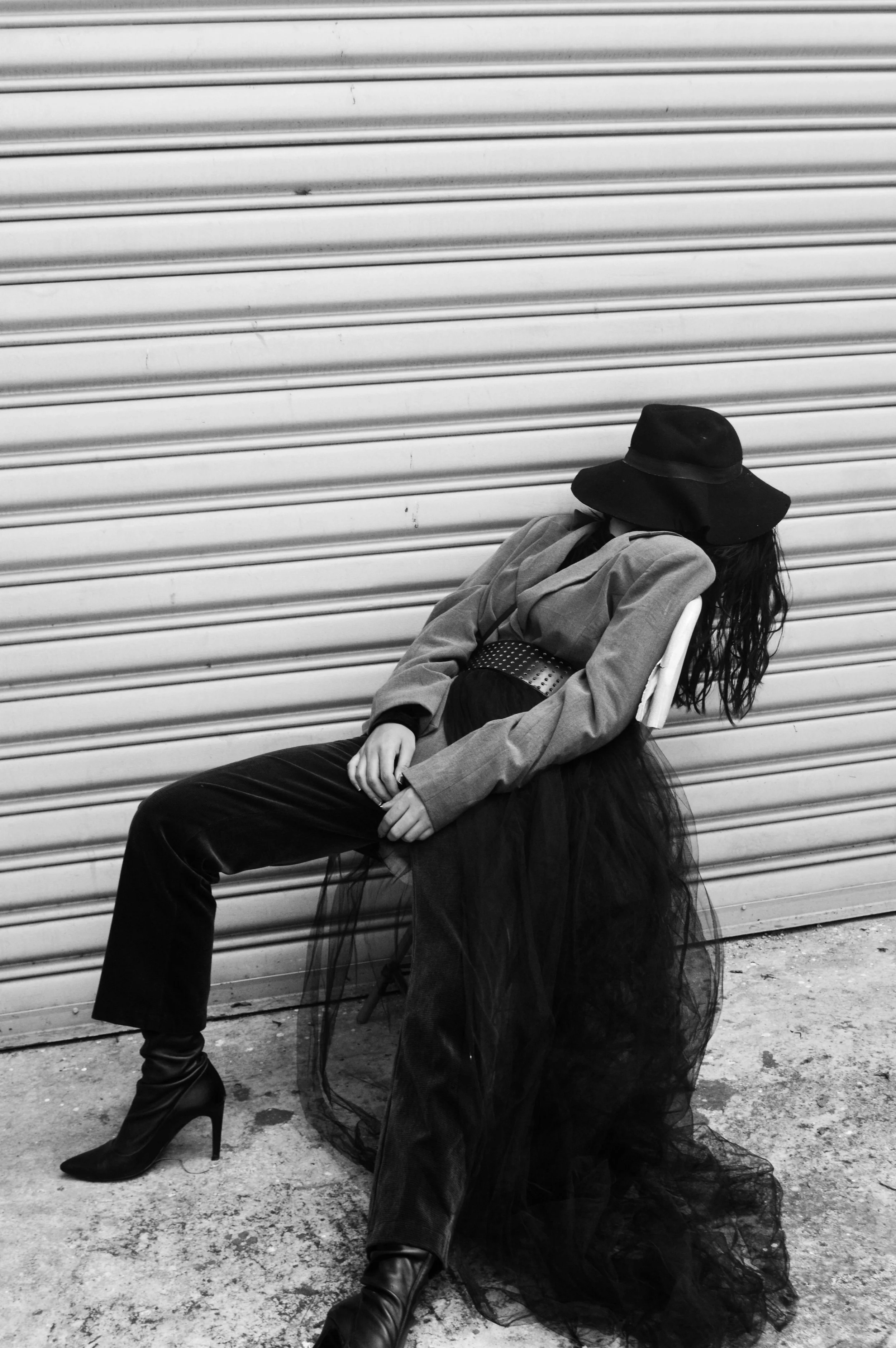 Black and white photo of a woman leaning back on a chair against a corrugated metal wall, wearing a wide-brim hat, blazer, tulle skirt, and high-heeled boots.