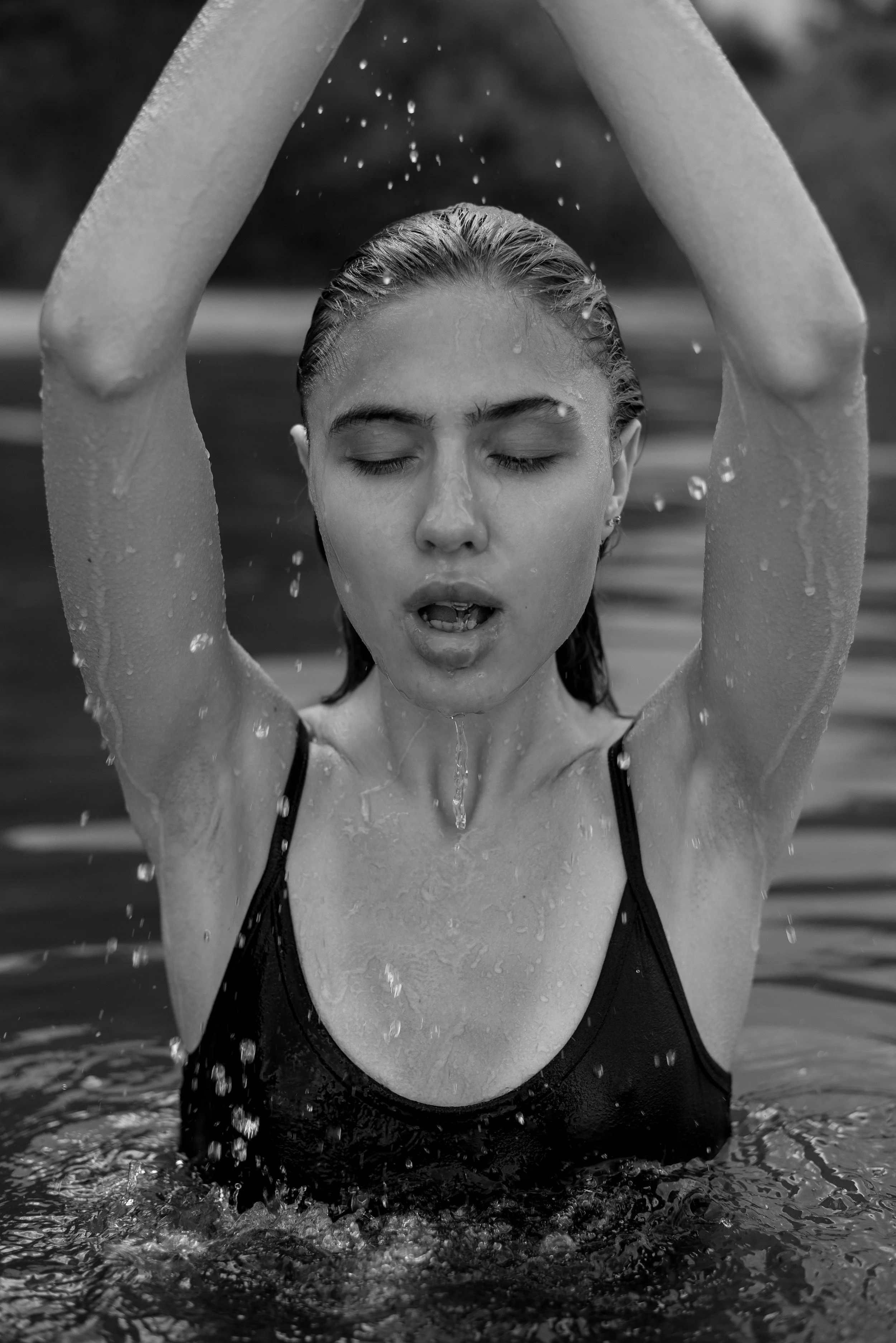 A woman with wet hair and closed eyes is emerging from the water, with her hands raised above her head, in a swimming pool.