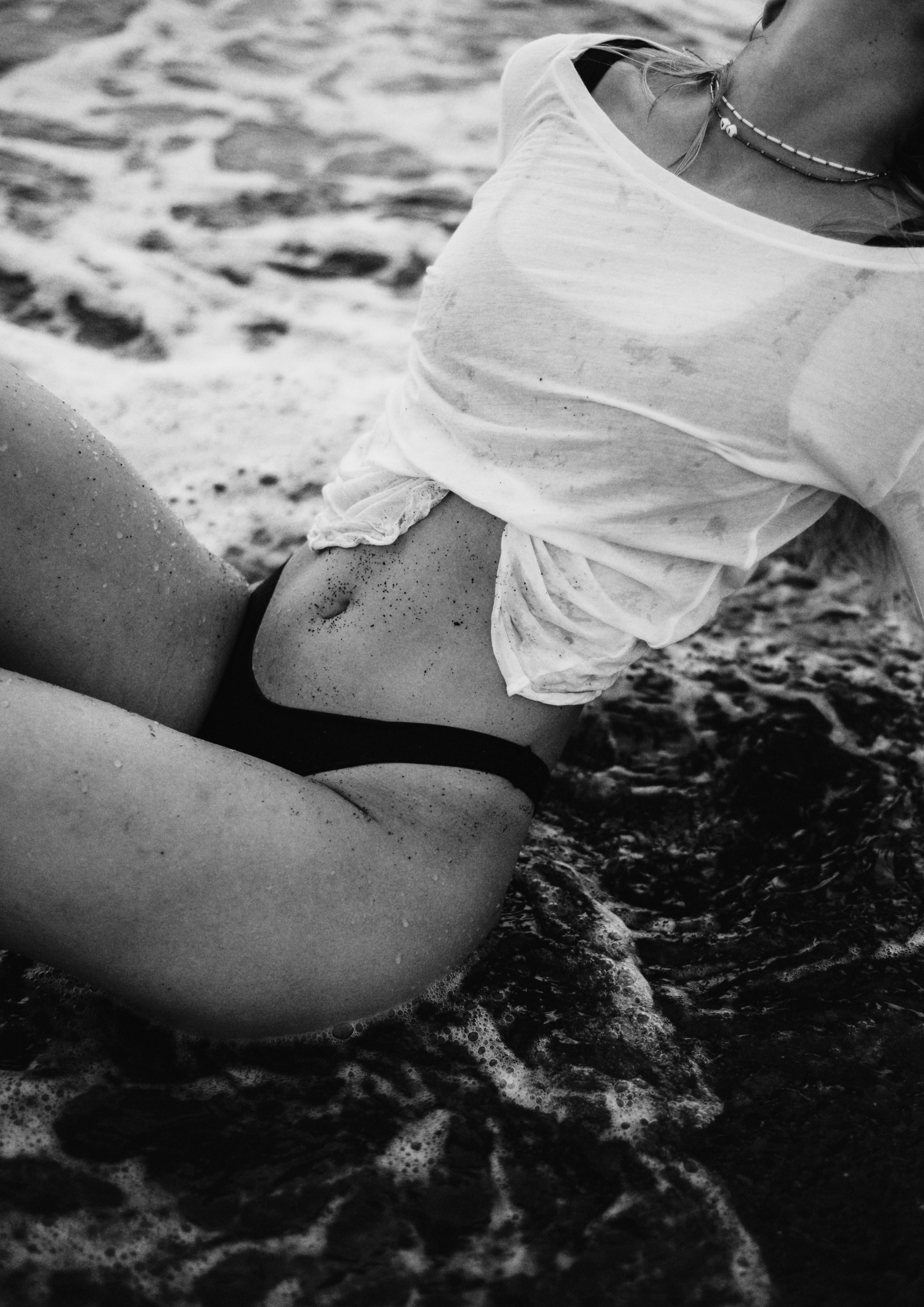 A black and white photo of a woman sitting on a sandy beach with water and foam around her. She is wearing a wet white shirt, black underwear, and a necklace.
