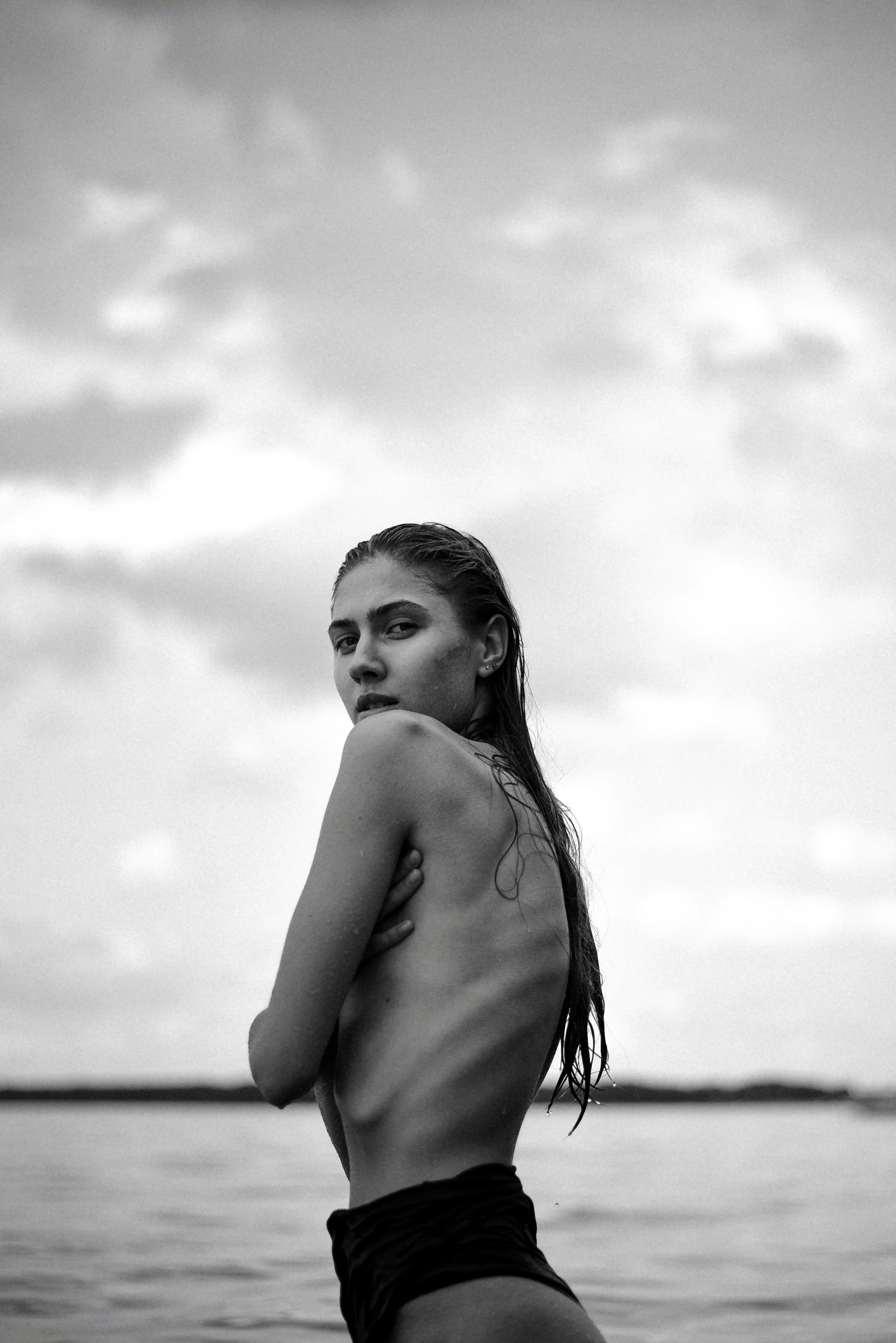 A black-and-white photograph of a young woman standing outdoors near water, with wet hair, wearing dark shorts, and looking over her shoulder at the camera.