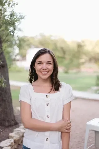 Young woman smiling outdoors next to a tree, wearing a white short-sleeve button-up shirt.