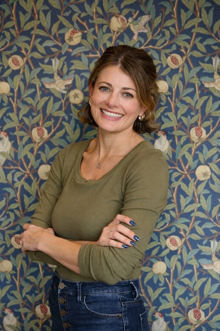 A woman with short brown hair smiling and standing with arms crossed in front of a floral patterned wallpaper.