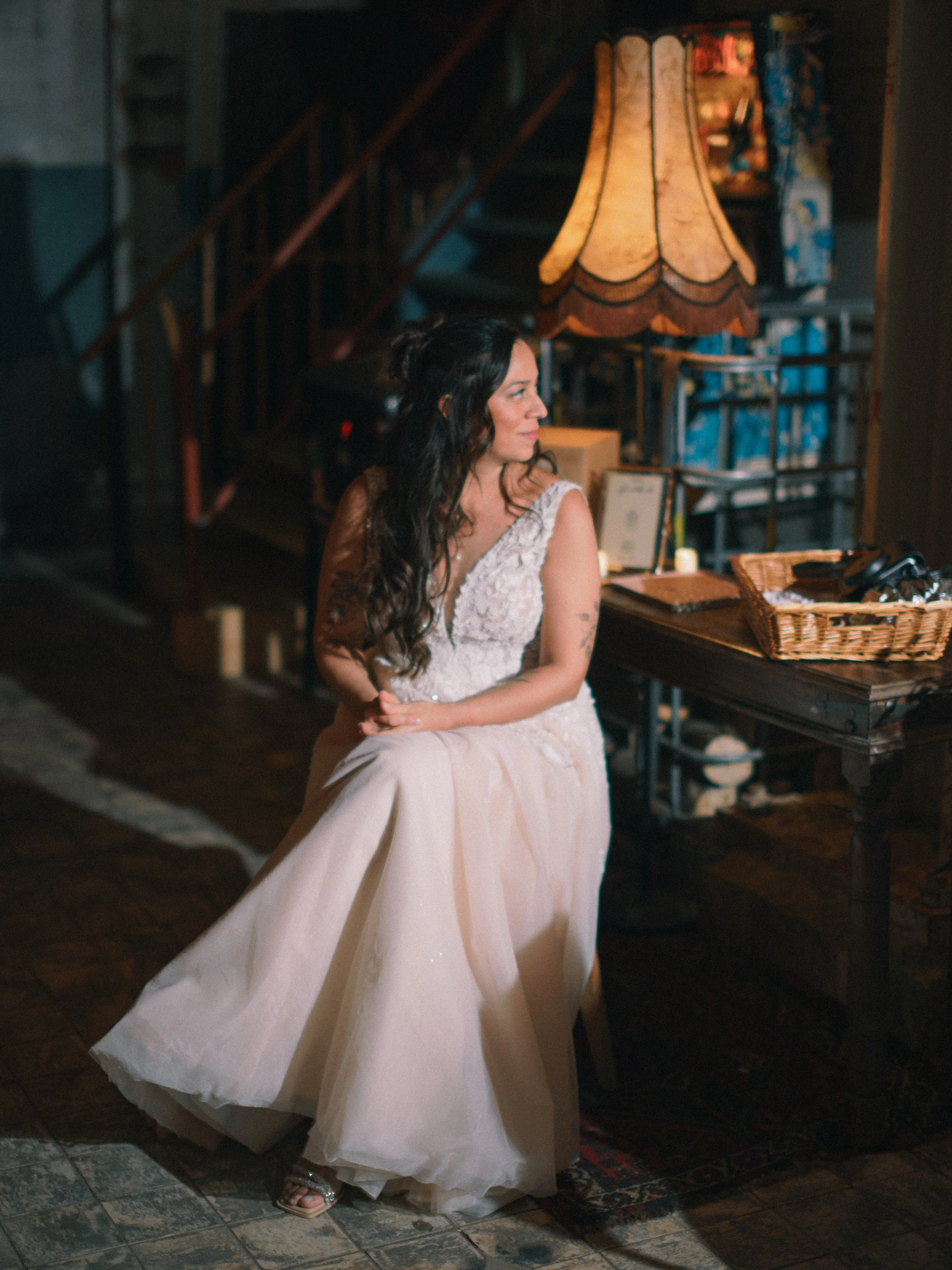 Vrouw in een lange witte jurk zit in een sfeervol vintage interieur met een grote lamp en manden.