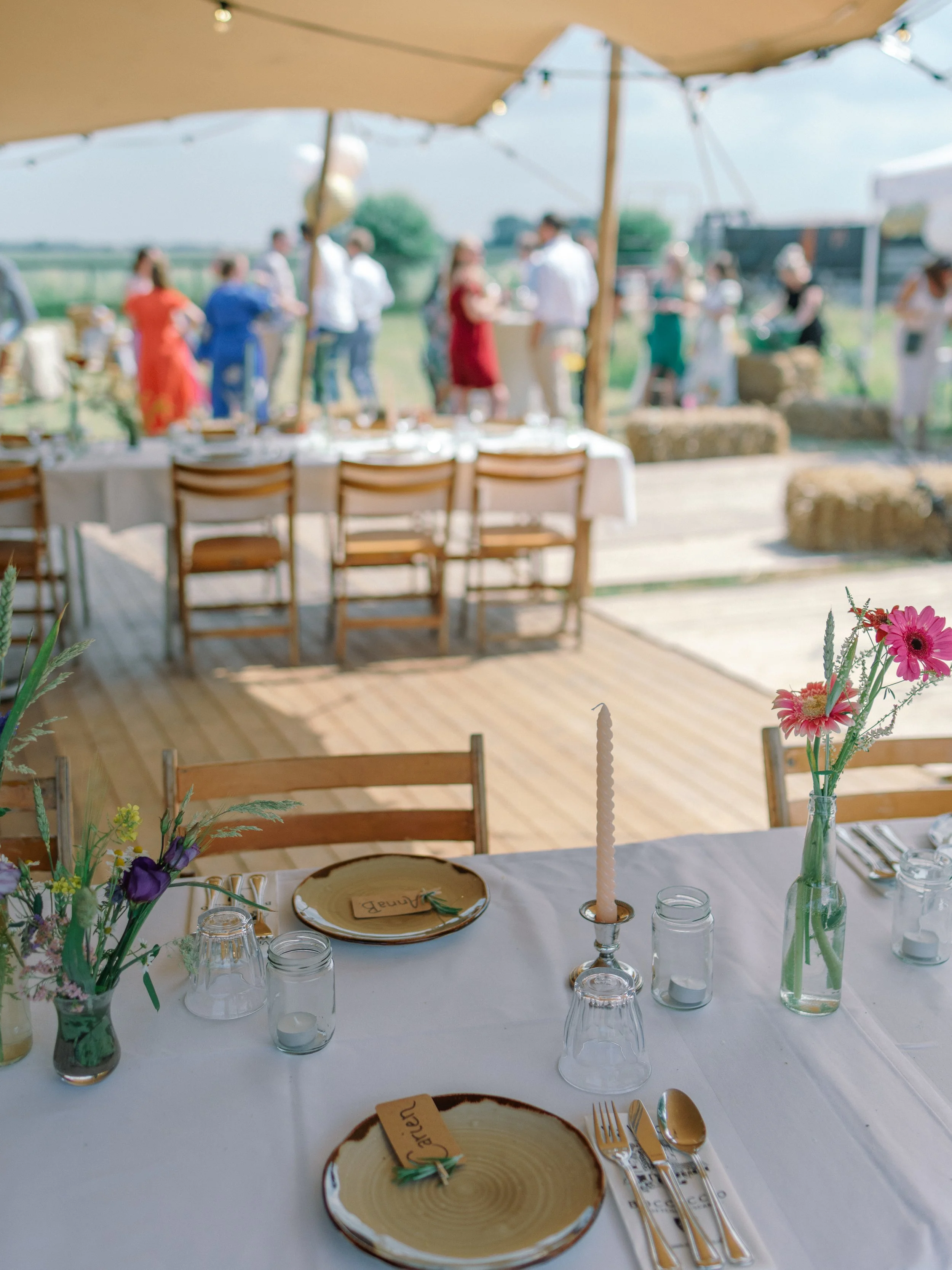 Feestelijk gedekte tafel onder een tent, met bloemen, kaarsen, servies en gasten op de achtergrond.