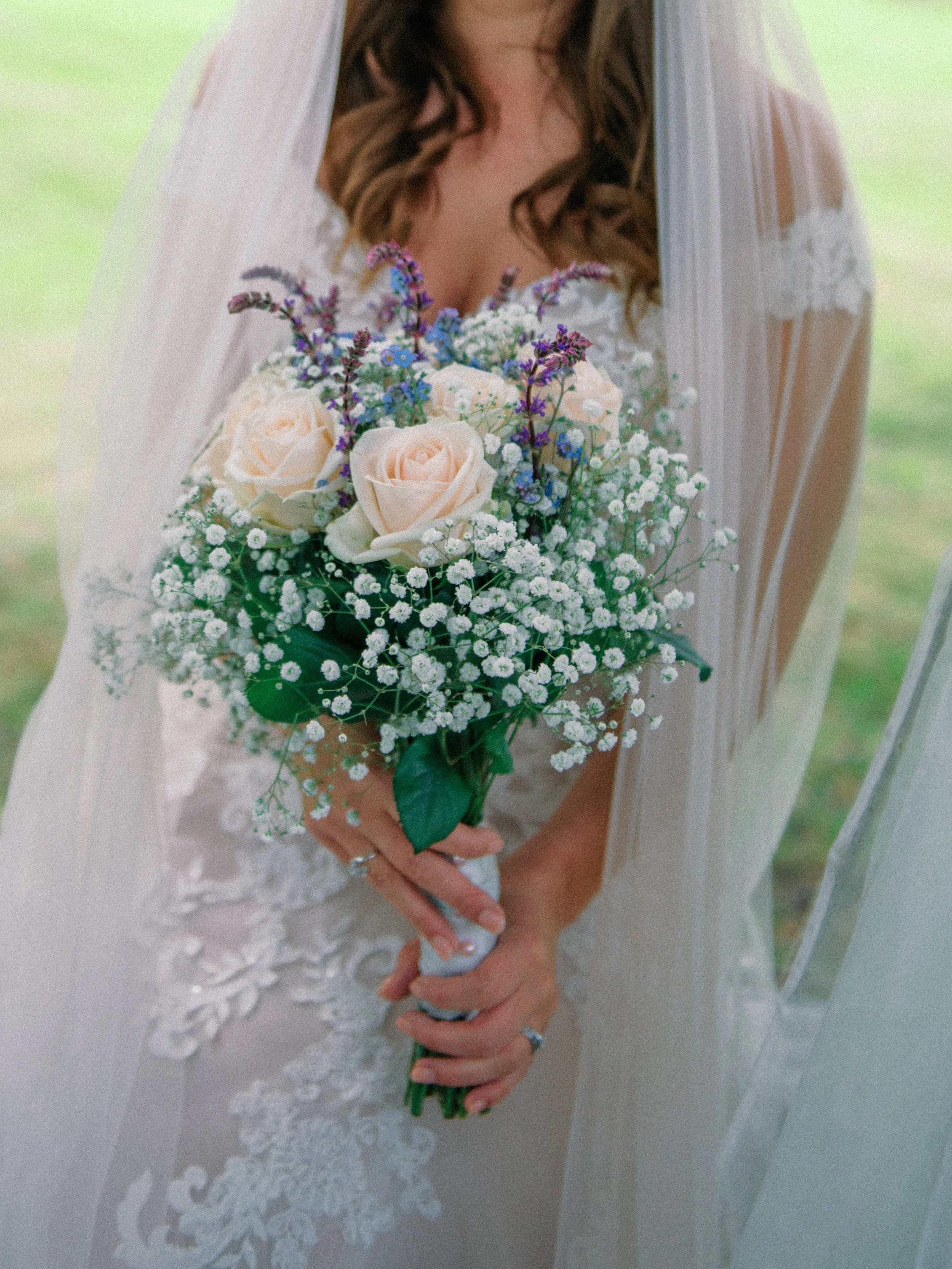 Bruidsportret van een bruid in witte sluier met een kleurrijk boeket van perzikkleurige rozen, blauwe bloemen en gipskruid.