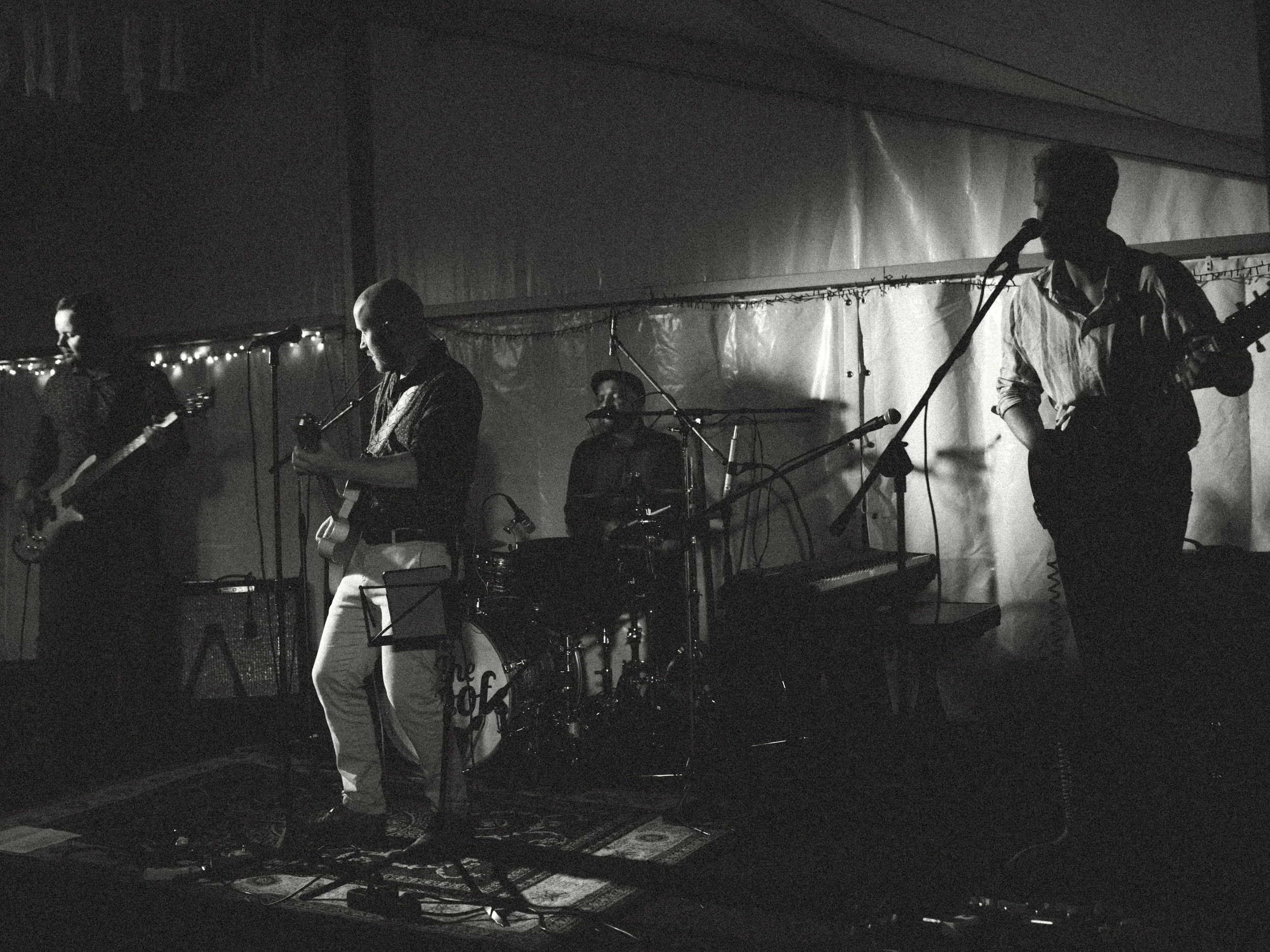 Zwart-witfoto van een vierkoppige band die live optreedt met gitaren, basgitaar en drums op een sfeervol verlicht podium.