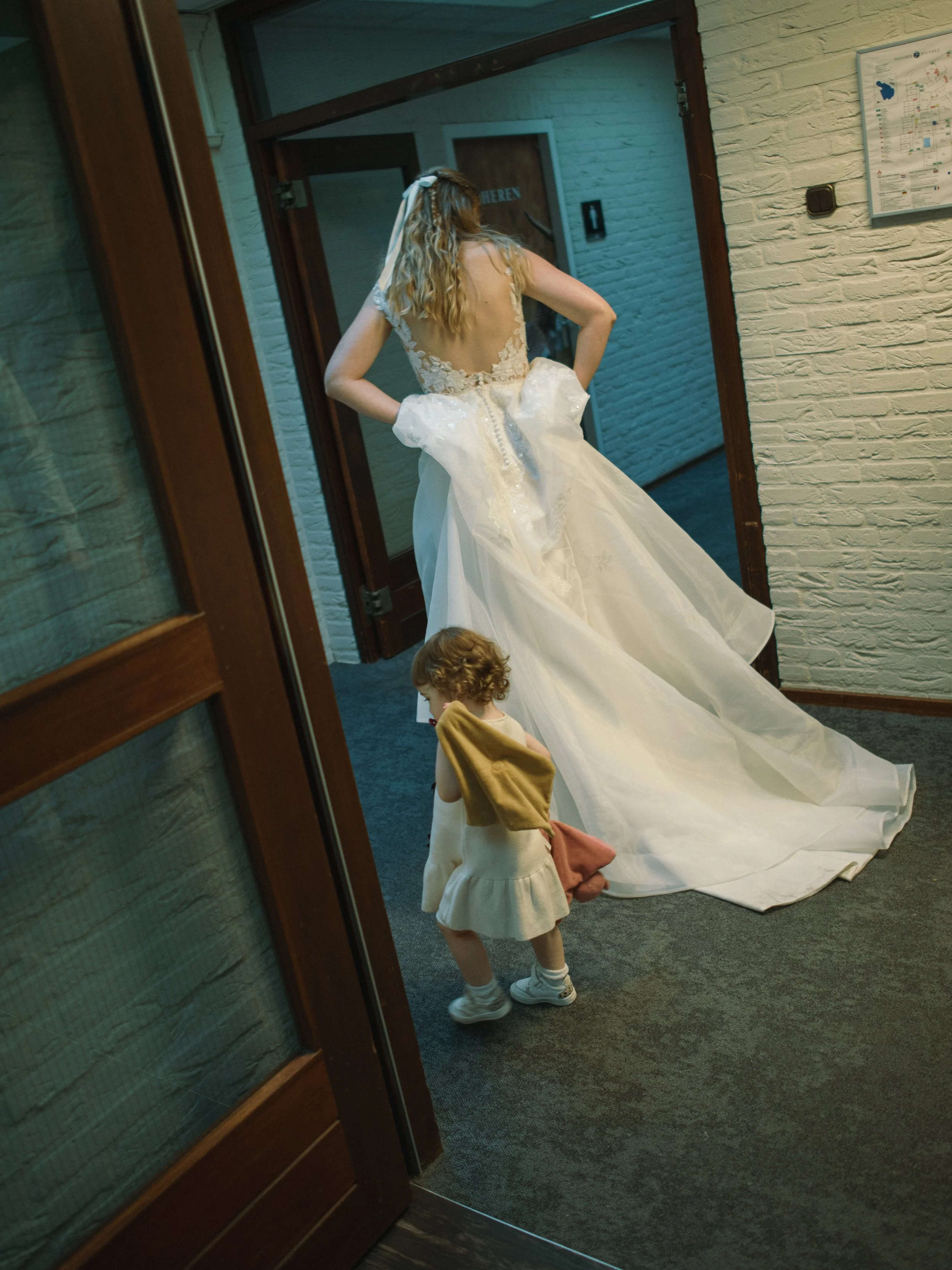 Bruid in een witte trouwjurk loopt door een deuropening, terwijl een klein kind haar sleep vasthoudt.