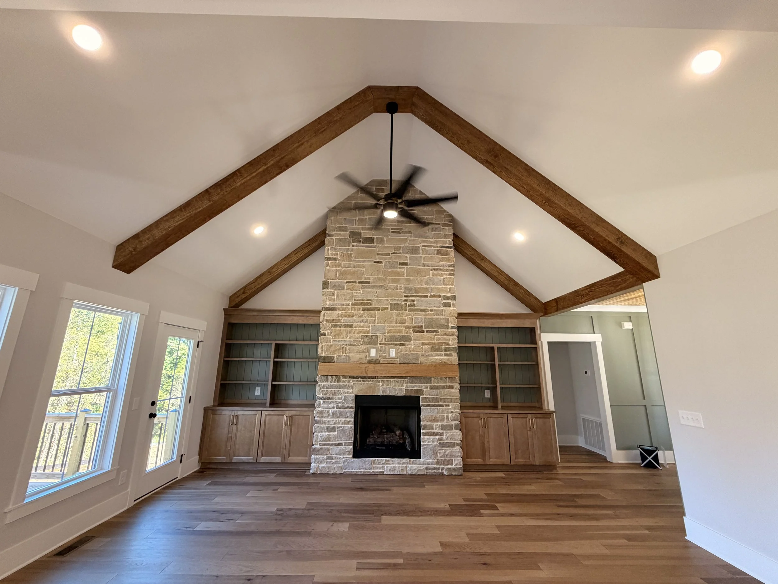 Living room with a vaulted ceiling, wooden beams, a stone fireplace, built-in shelves, hardwood flooring, and large windows with a view of the outside.
