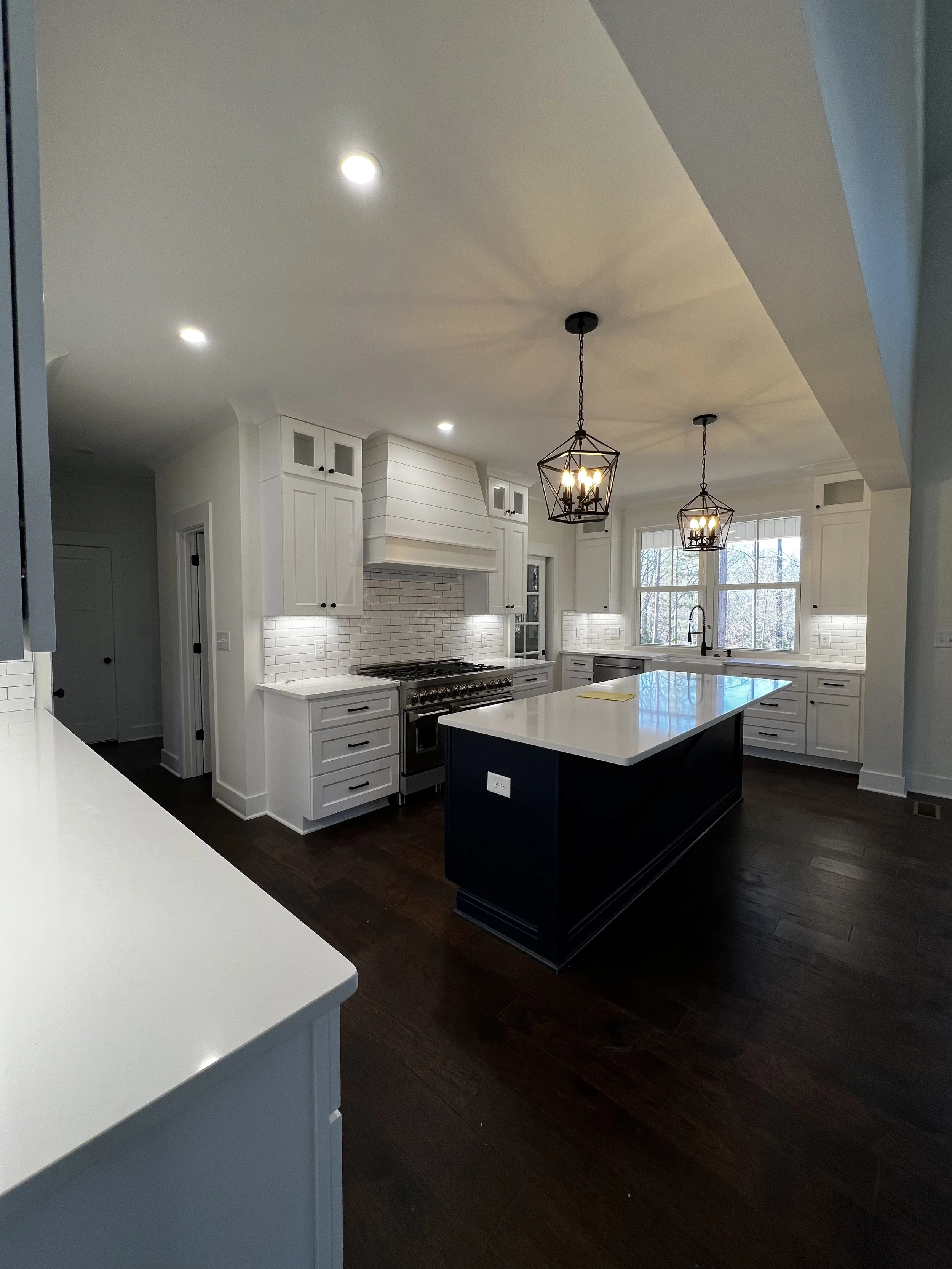 Modern kitchen with white cabinets, a large island with a white countertop, dark hardwood floors, pendant lights hanging from the ceiling, large window, and brick backsplash beneath the vent hood.