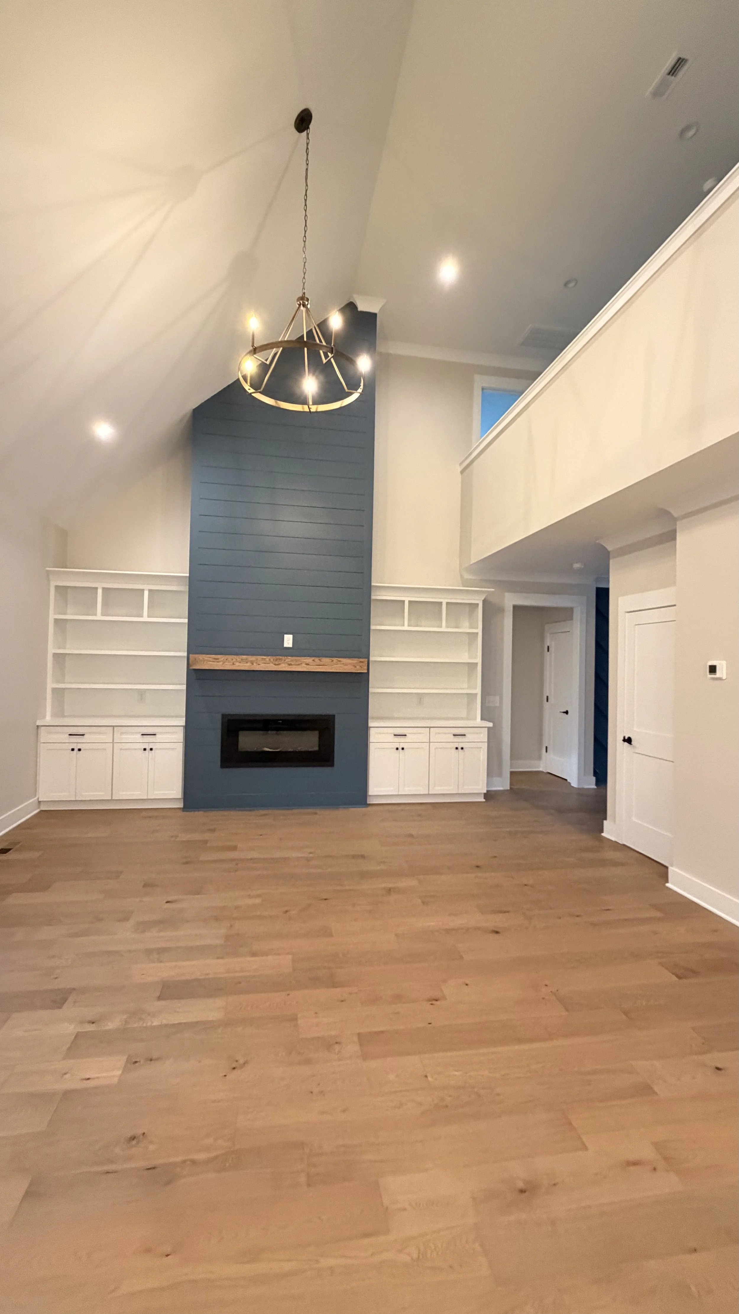 Living room with hardwood floors, white built-in shelves, a fireplace with a blue accent wall, a black chandelier, and a high ceiling with a balcony.