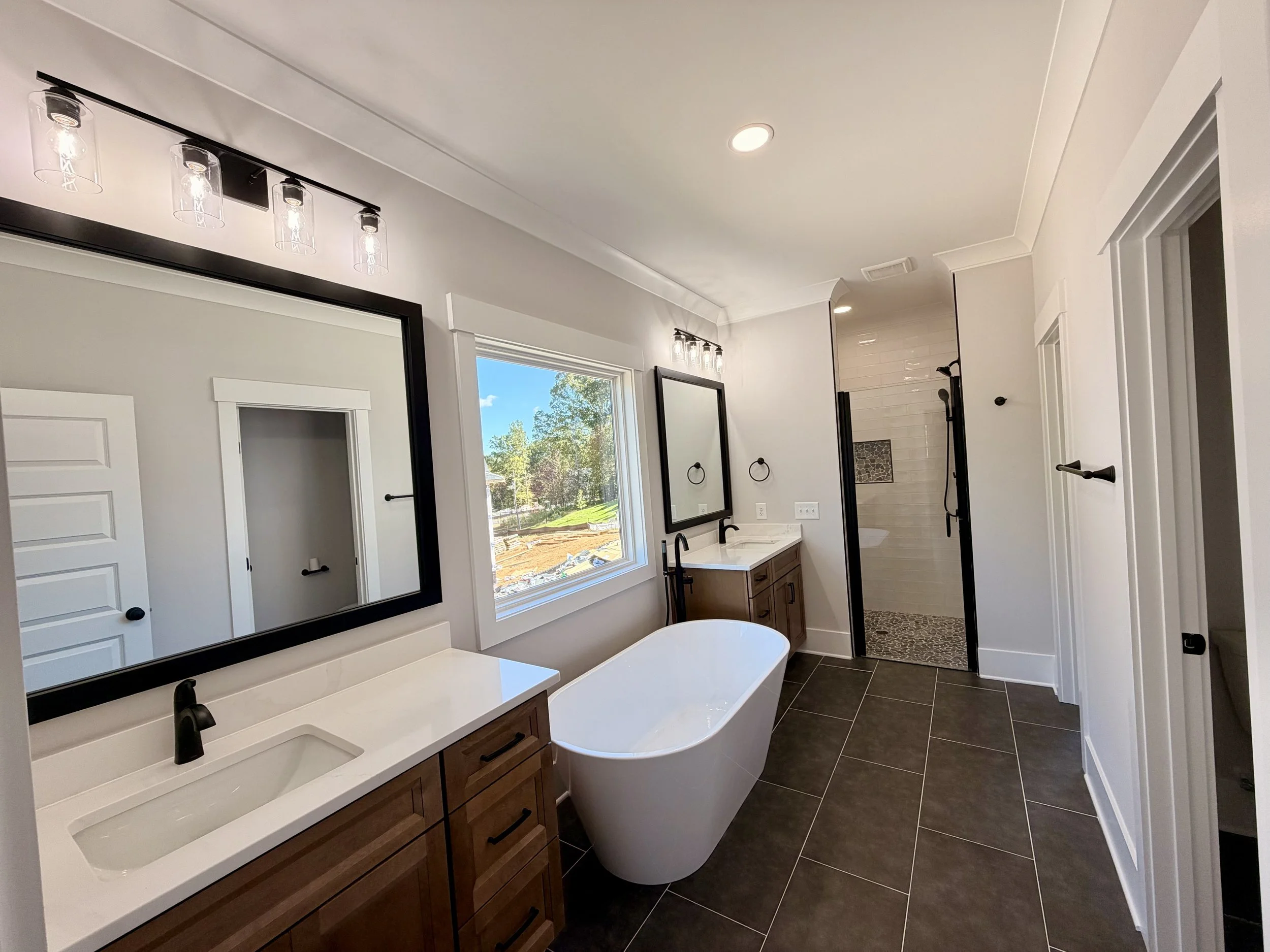 Modern bathroom with a white tub, vanity with double sink, large mirror, black fixtures, and a walk-in shower with a glass door and white subway tile walls.