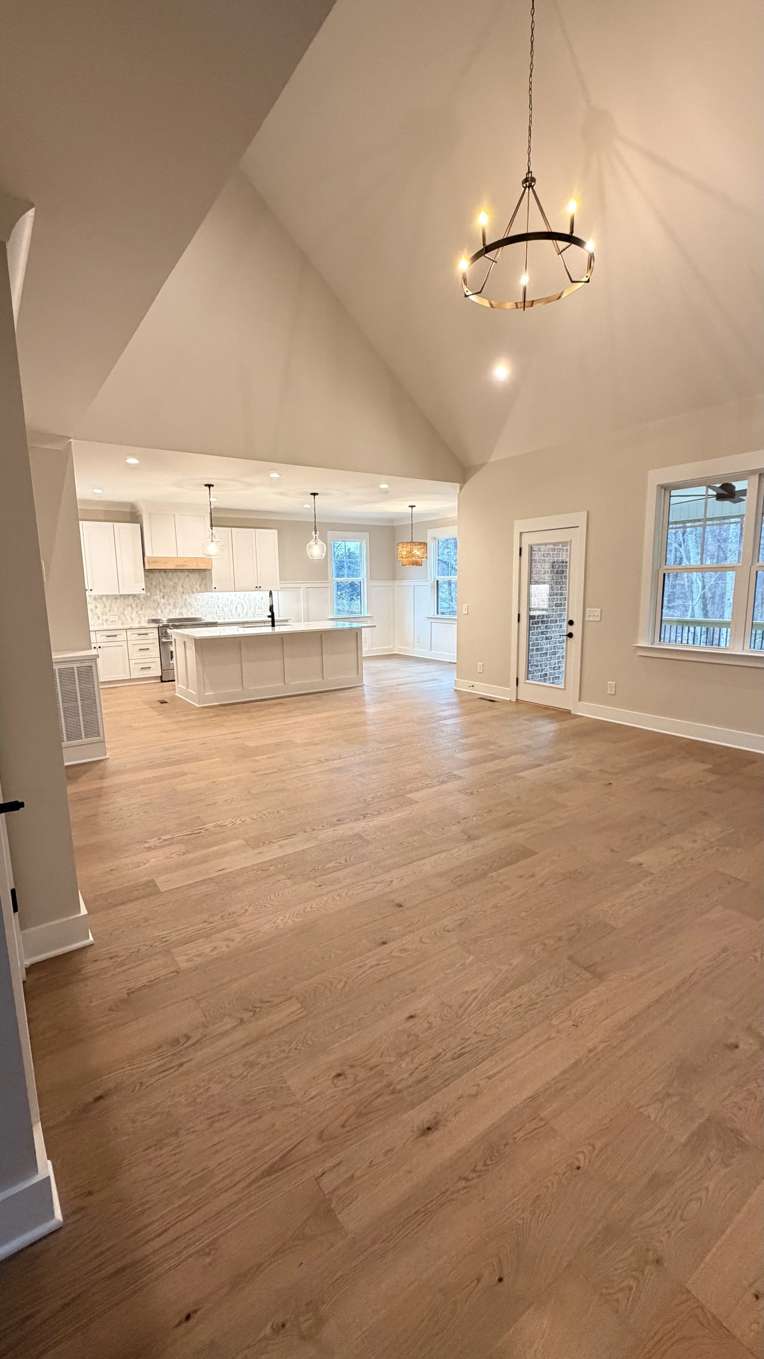 Open-concept living area with hardwood floors, a view of a kitchen with white cabinets, a large island, pendant lighting, and windows showing a wooded outdoor scene.