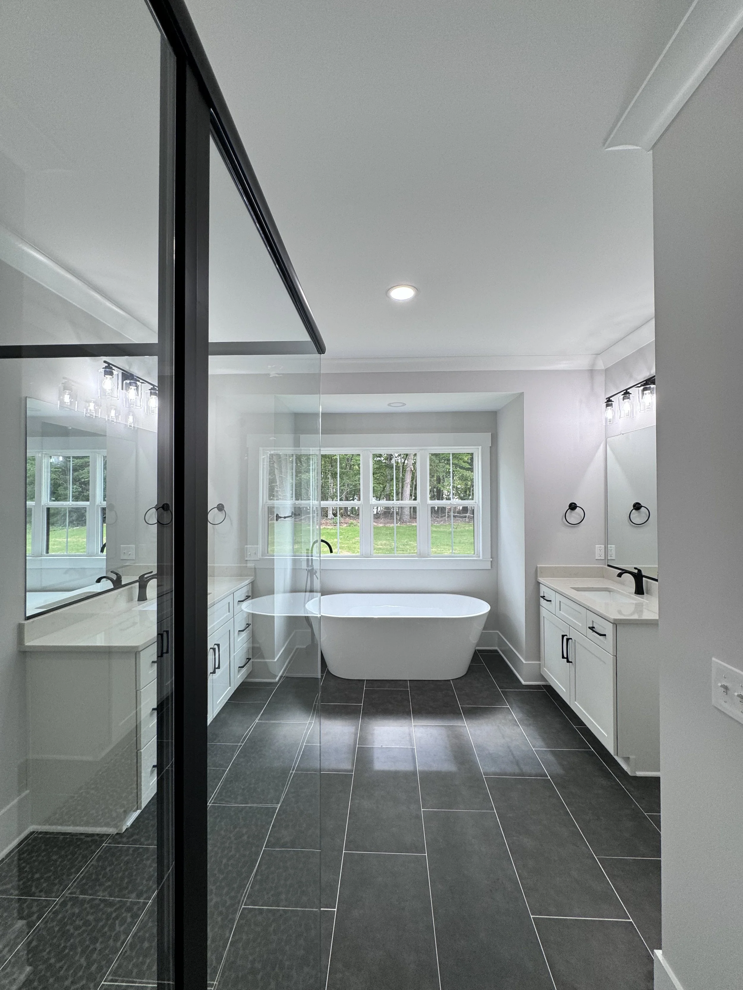 Modern bathroom with a freestanding bathtub, large windows, white cabinetry, double sinks, black fixtures, and dark floor tiles.