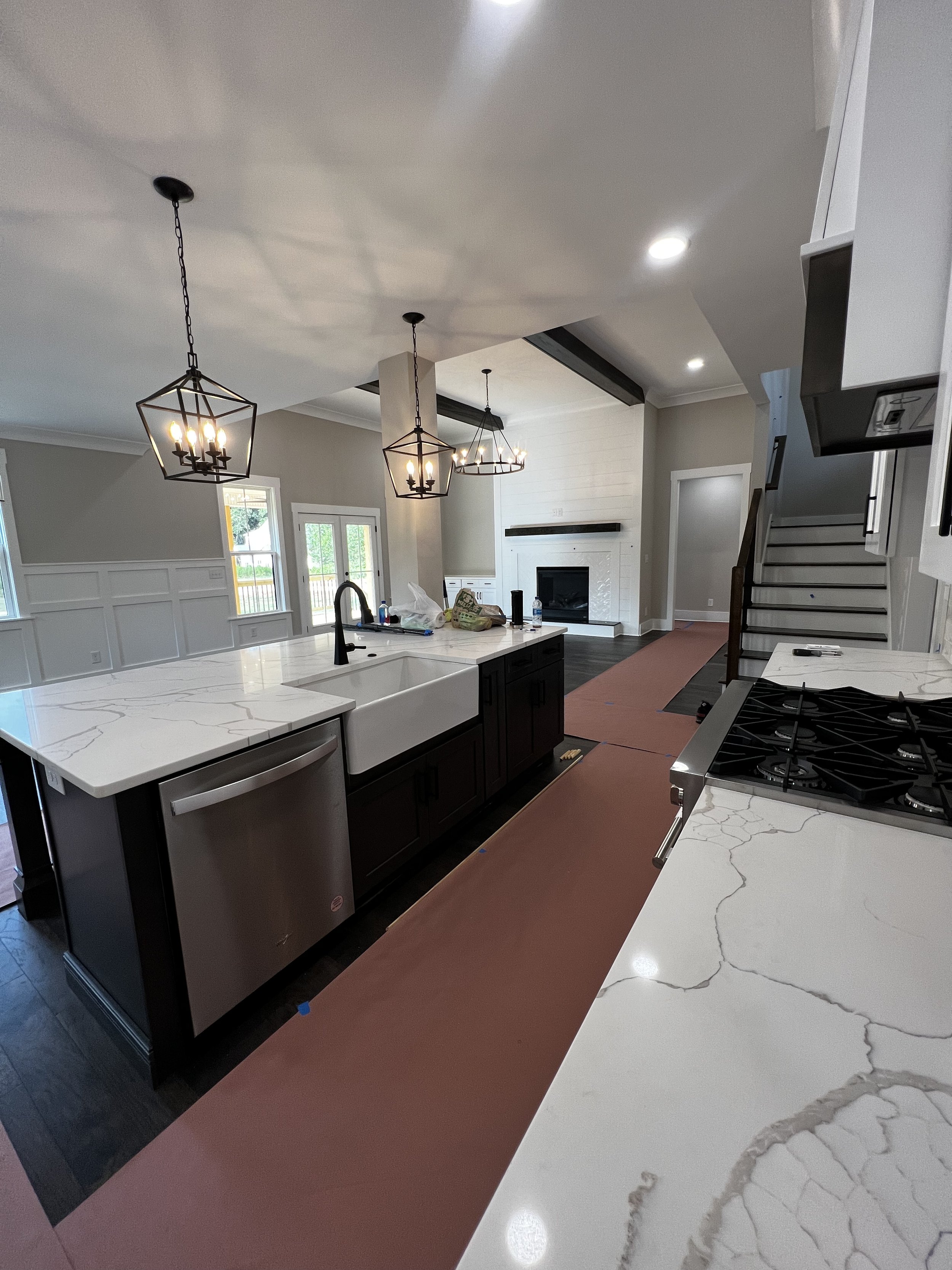 Modern kitchen under renovation with white marble countertops, dark cabinetry, and pendant lights over the island, and a fireplace in the living room area.