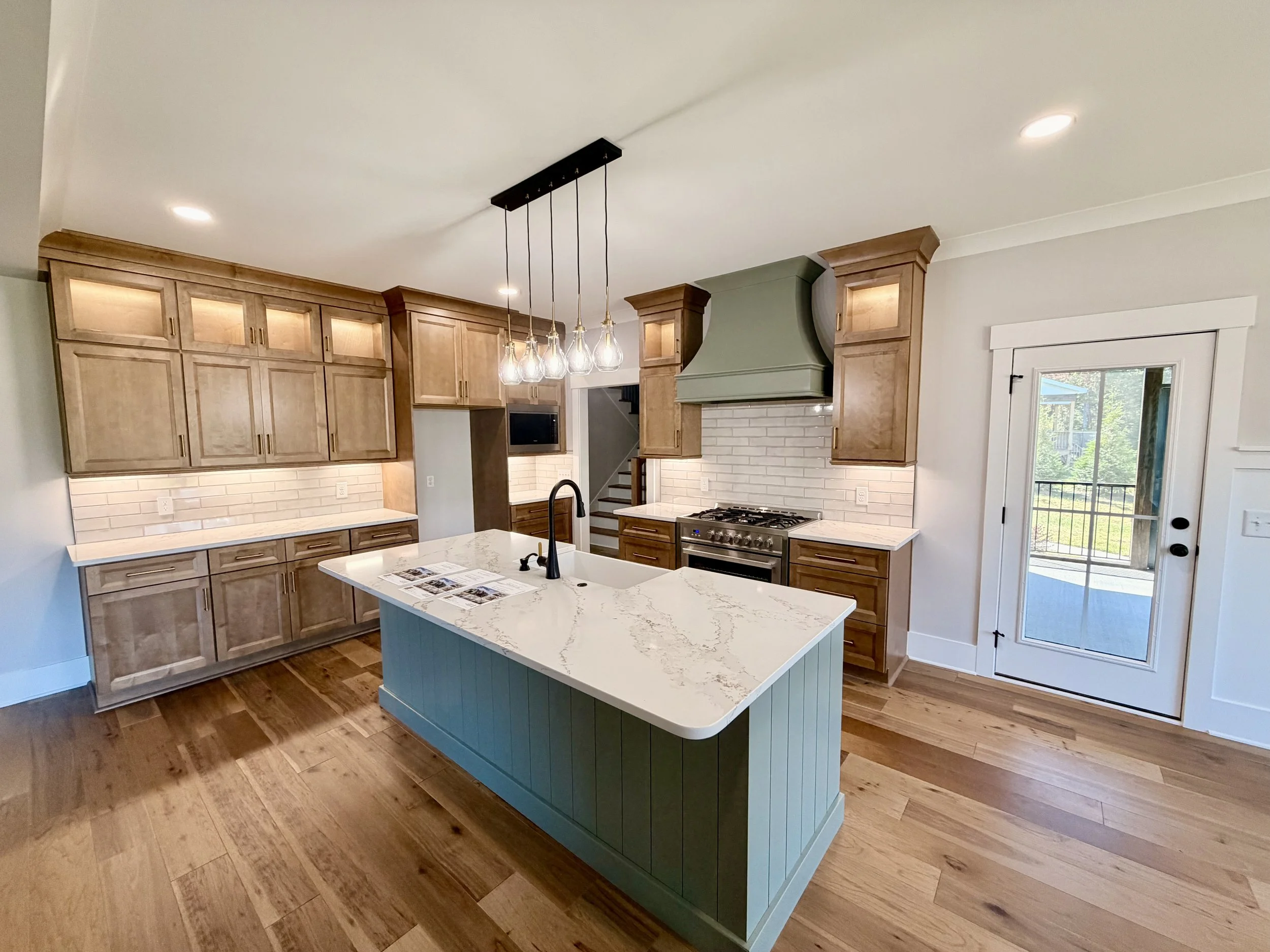 Modern kitchen featuring light wood cabinets, a green range hood, white subway tile backsplash, a white kitchen island with a marble countertop, pendant lights, and a sliding glass door leading outside.