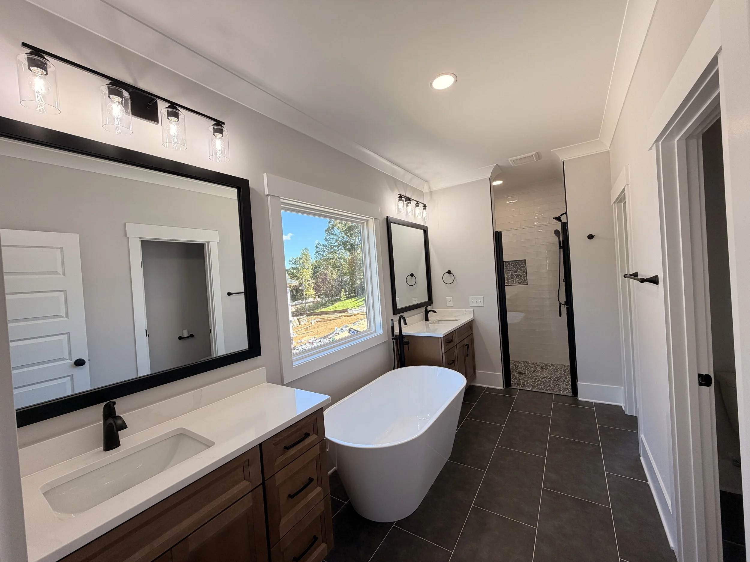 Modern bathroom with a large window, freestanding bathtub, double sink vanity with wooden cabinets, and walk-in shower with glass door and tiled interior.