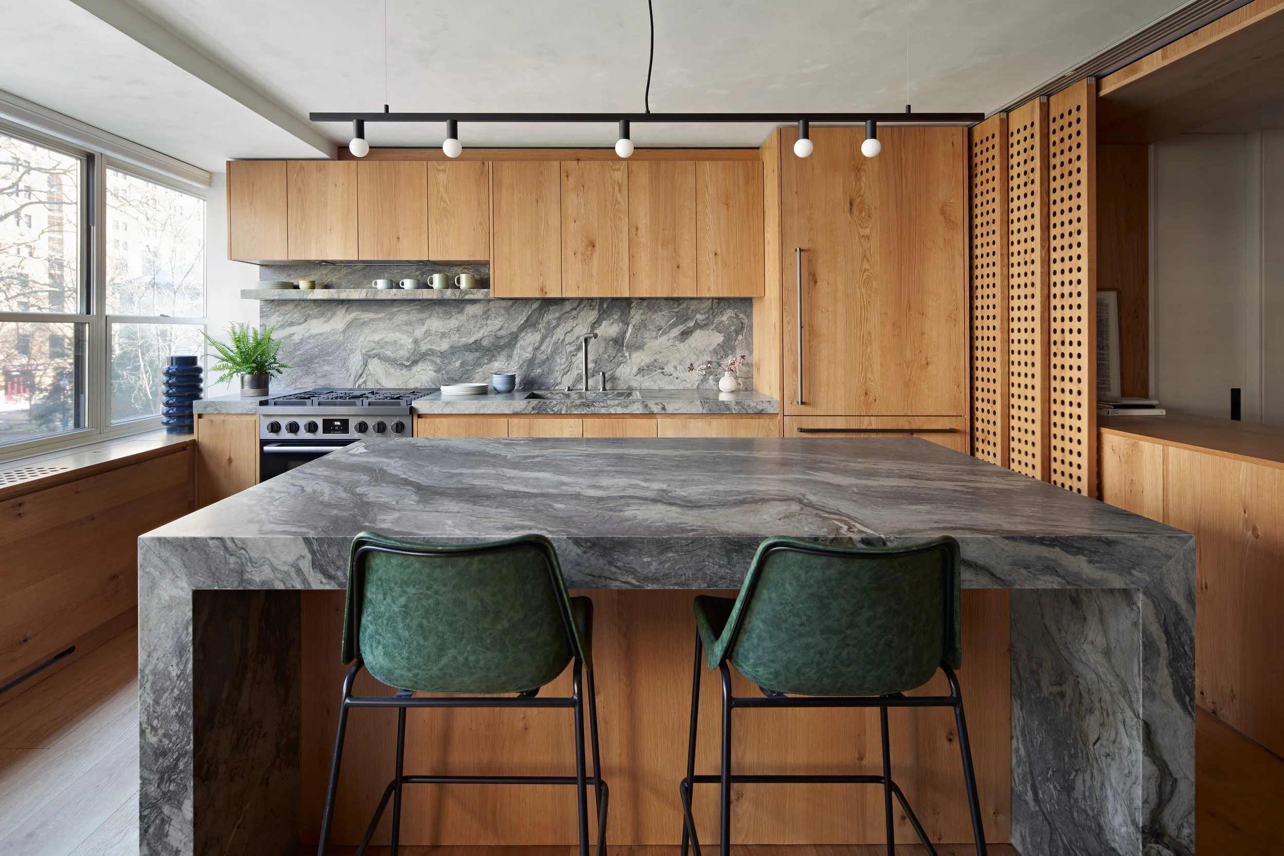 Modern kitchen with wooden cabinets, a marble backsplash and island, two green chairs, and large windows.
