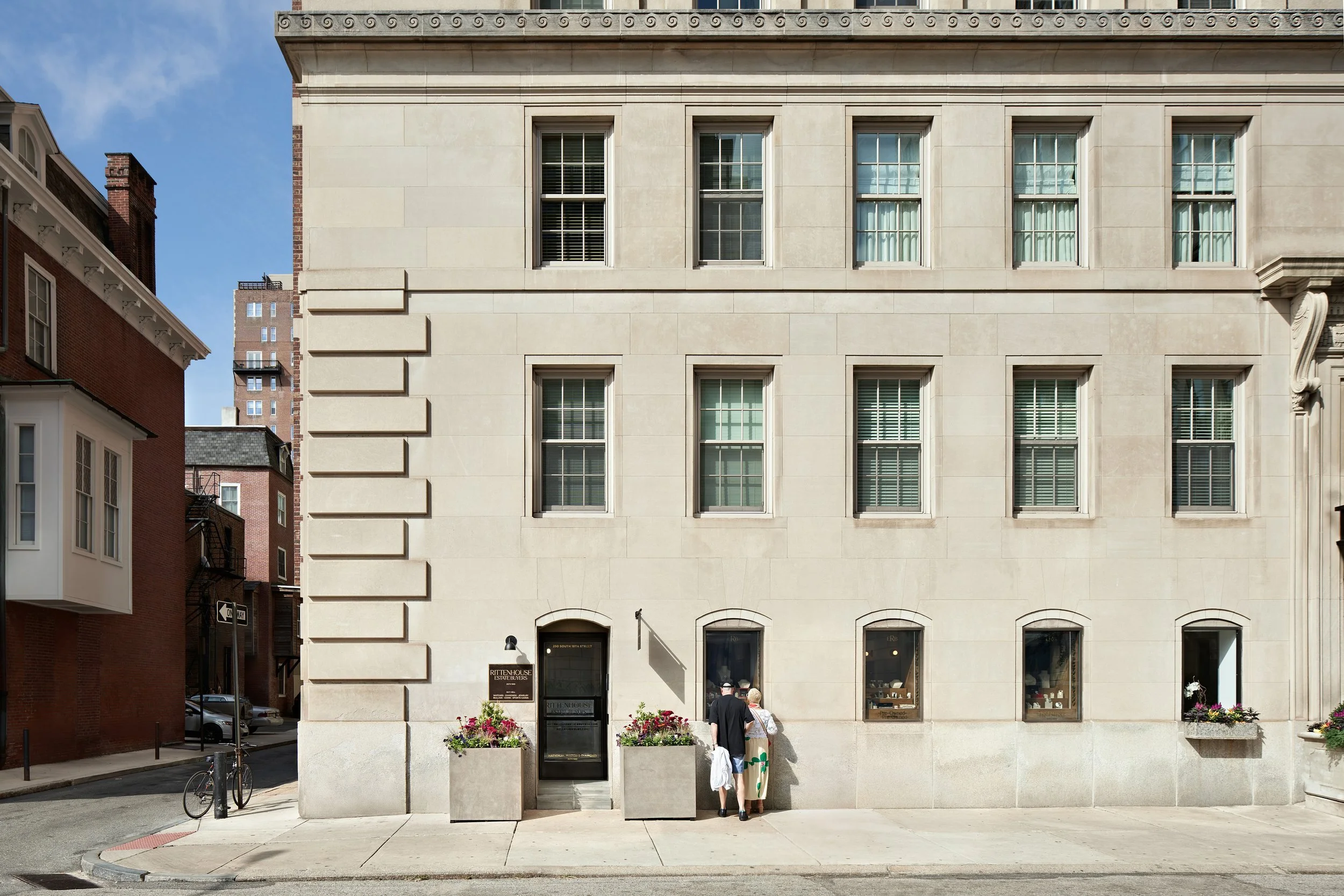 A beige stone building with four floors and six windows, two planters with flowers, and two people standing outside near the entrance