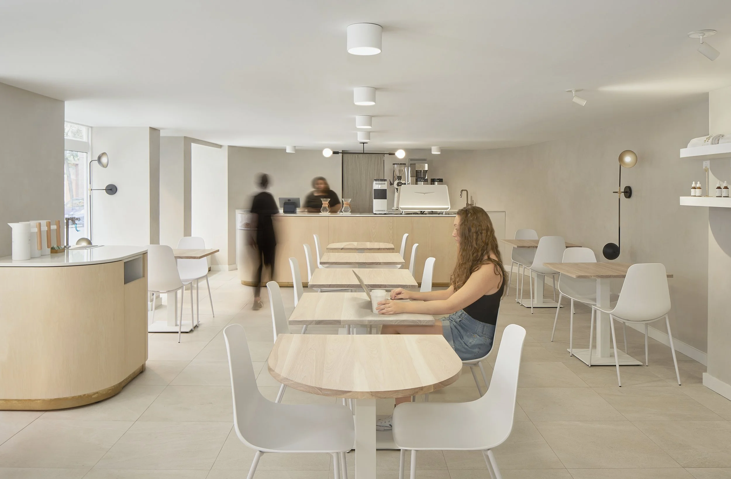 A woman sitting alone at a table in a modern, minimalistic cafe, with a latte in front of her, while two baristas work behind the counter. The cafe has light-colored wood furniture and neutral walls with sleek lighting.