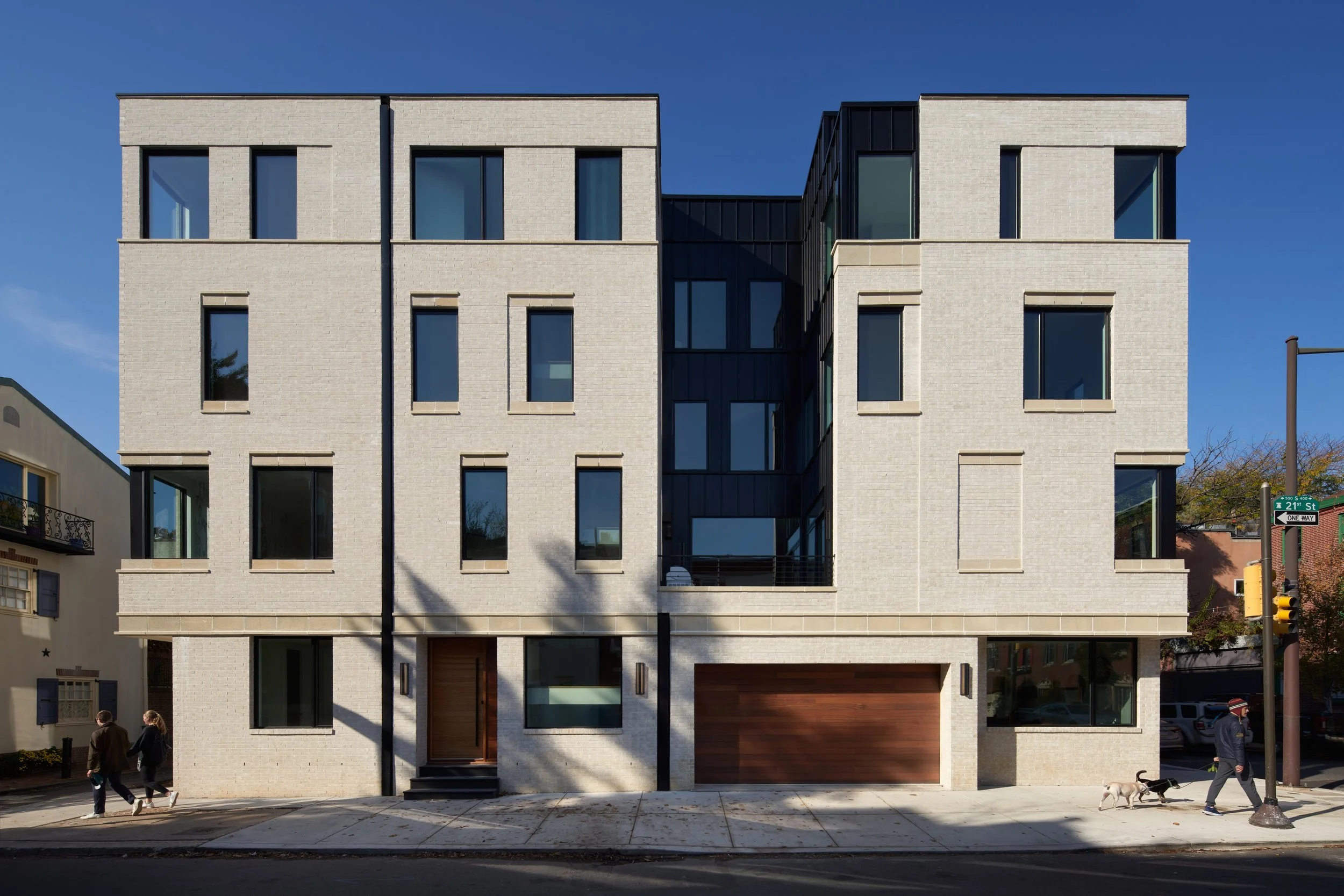 Modern multi-story residential building with white brick exterior and black window frames on a city street. People walking dogs and pedestrians near the sidewalk.