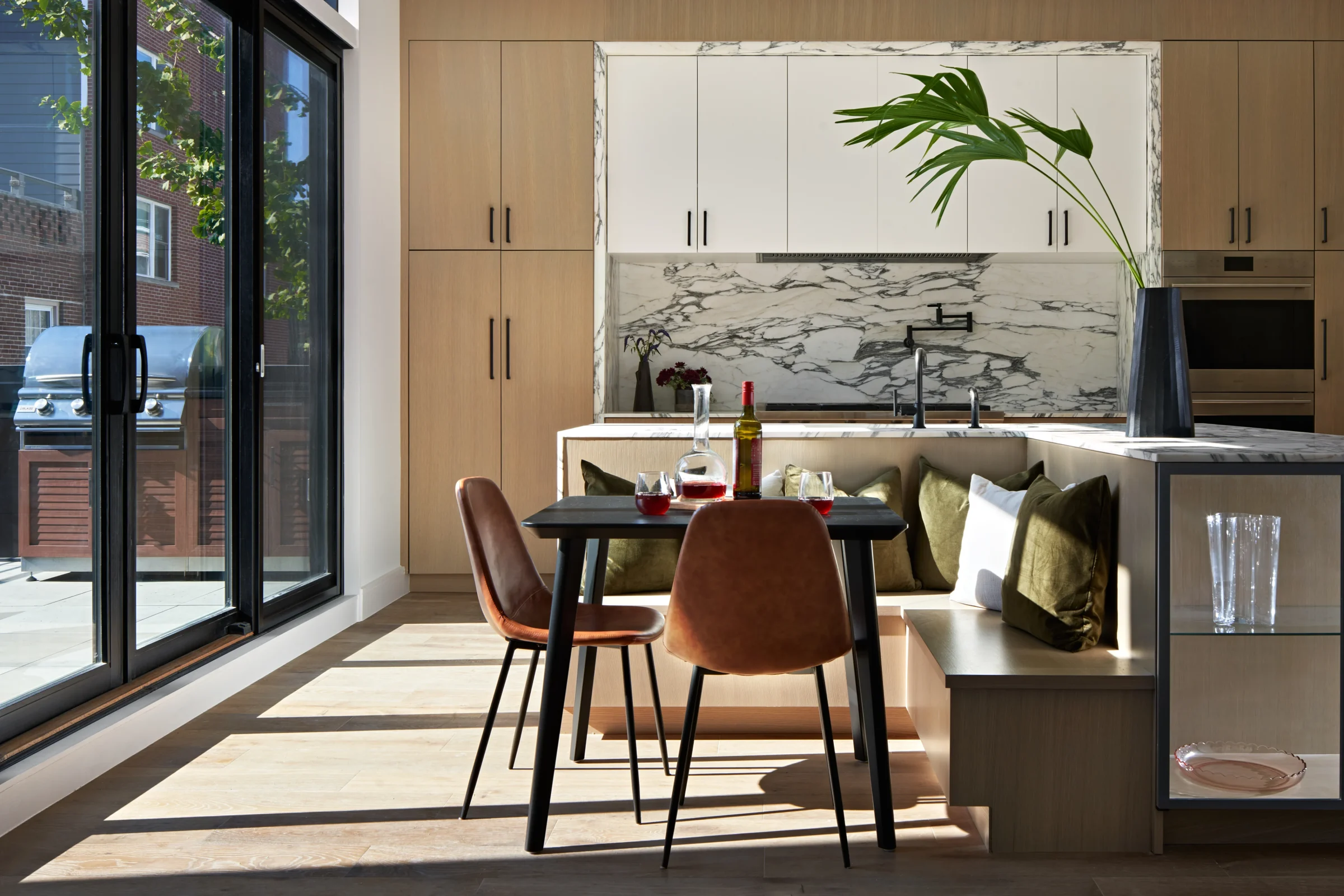 Modern kitchen with wooden and marble finishes, sliding glass door with outdoor grill, dining table with wine and glasses, and large potted plant.