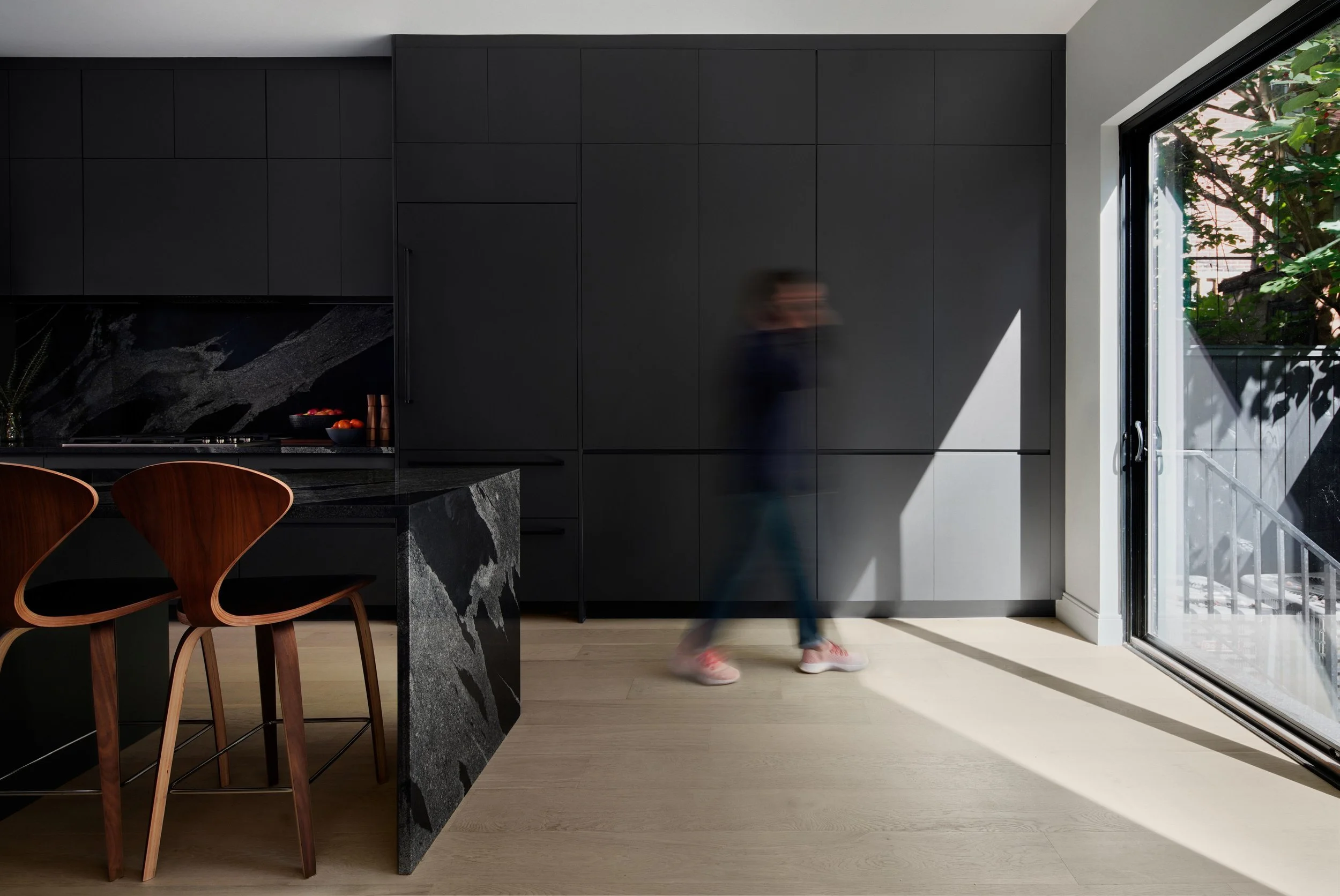 Modern kitchen with black cabinets, a marble island, wooden chairs, and a large glass door showing outdoor greenery. A blurred person is walking inside.