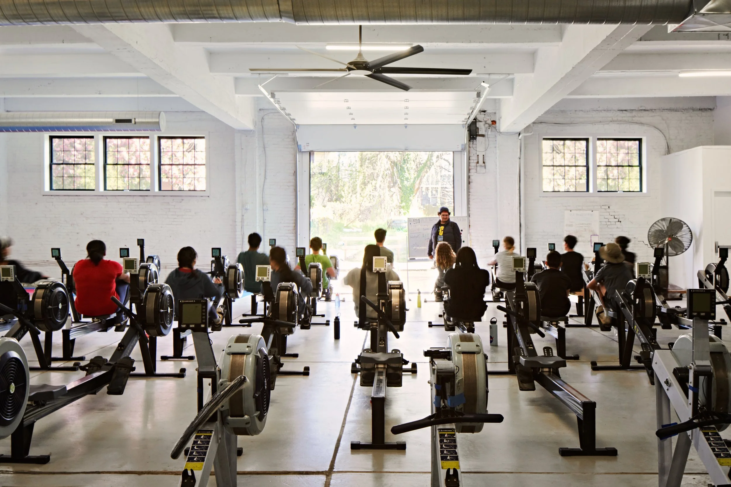 Indoor rowing class with multiple rowing machines and a teacher at the front leading students in a spacious studio with large windows and an open garage door.
