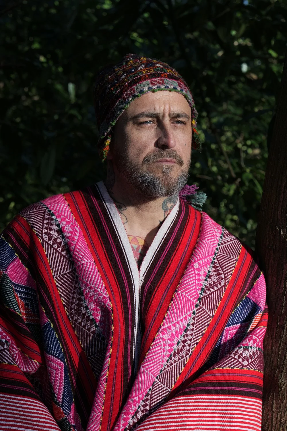 A man with a beard and mustache wearing a colorful, patterned blanket and a multicolored knit hat, standing outdoors with a dark, leafy background.