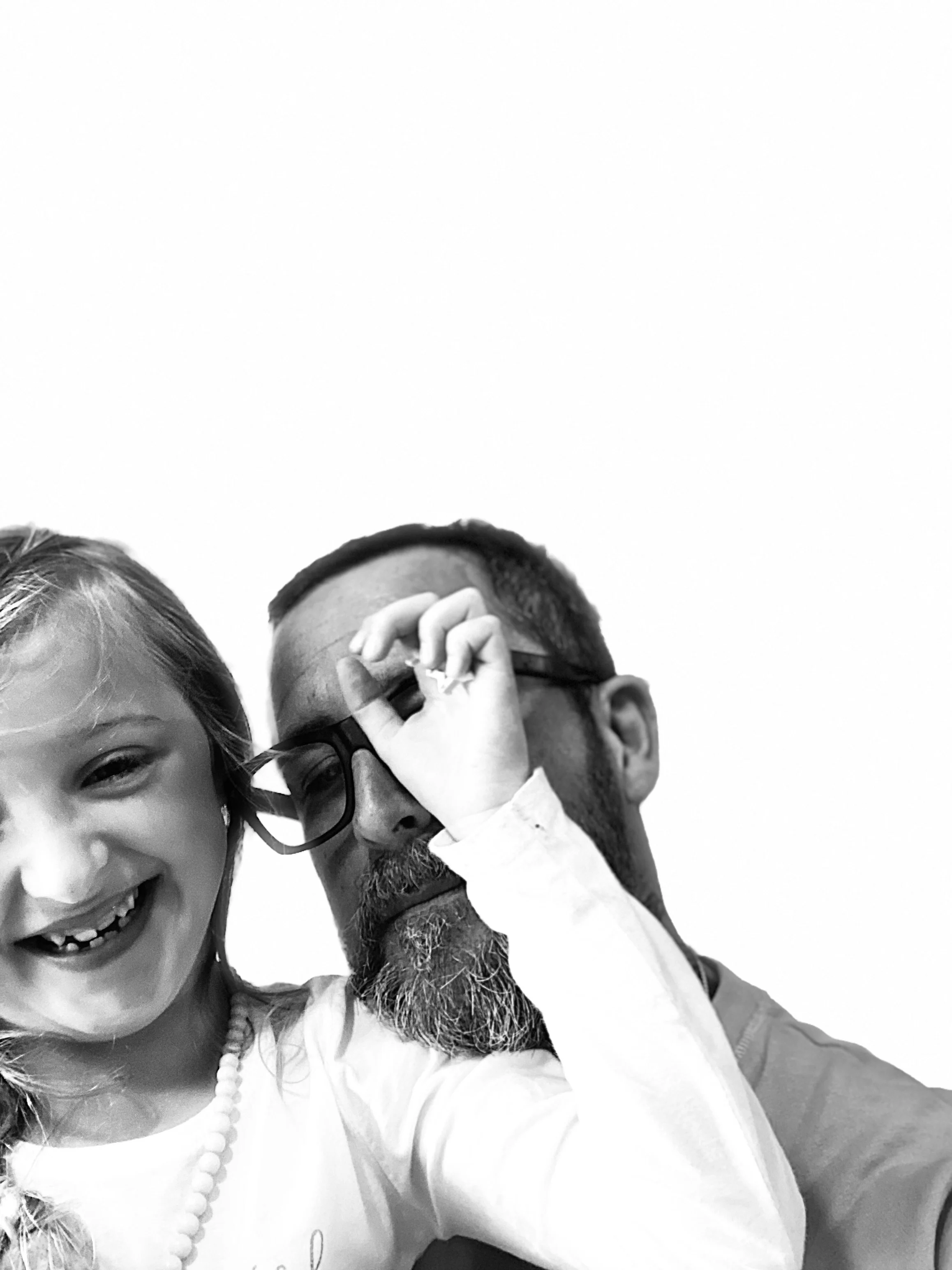 A smiling young girl with curly hair, wearing earrings and a necklace, taking a selfie with a man wearing glasses and a beard, who is adjusting his glasses.