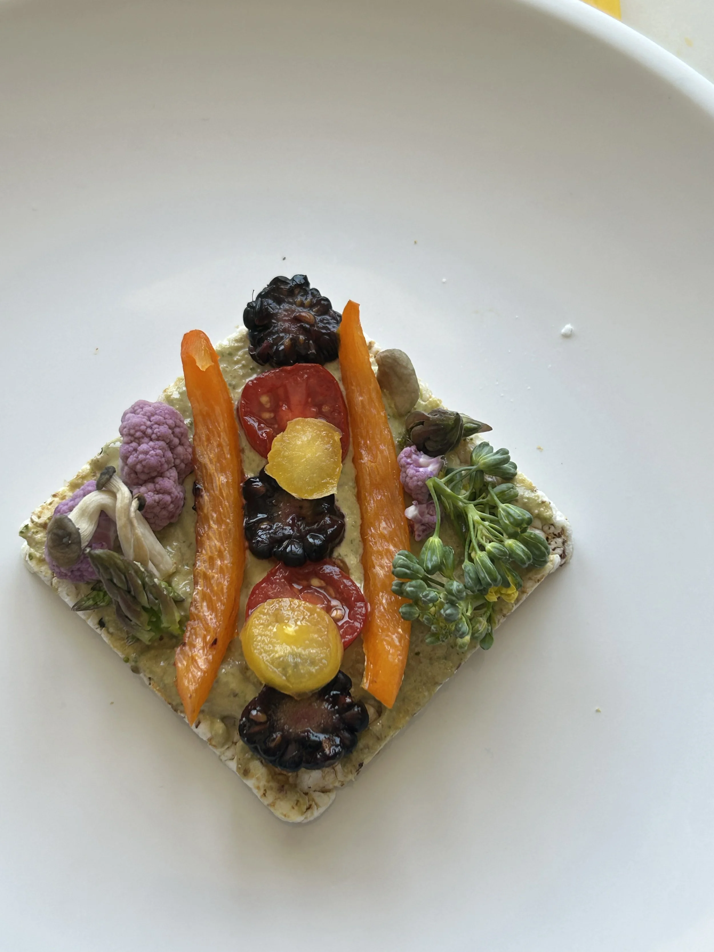 A plate of assorted vegetables including carrots, cherry tomatoes, blackberries, purple cauliflower, and green sprouts on a square cracker.