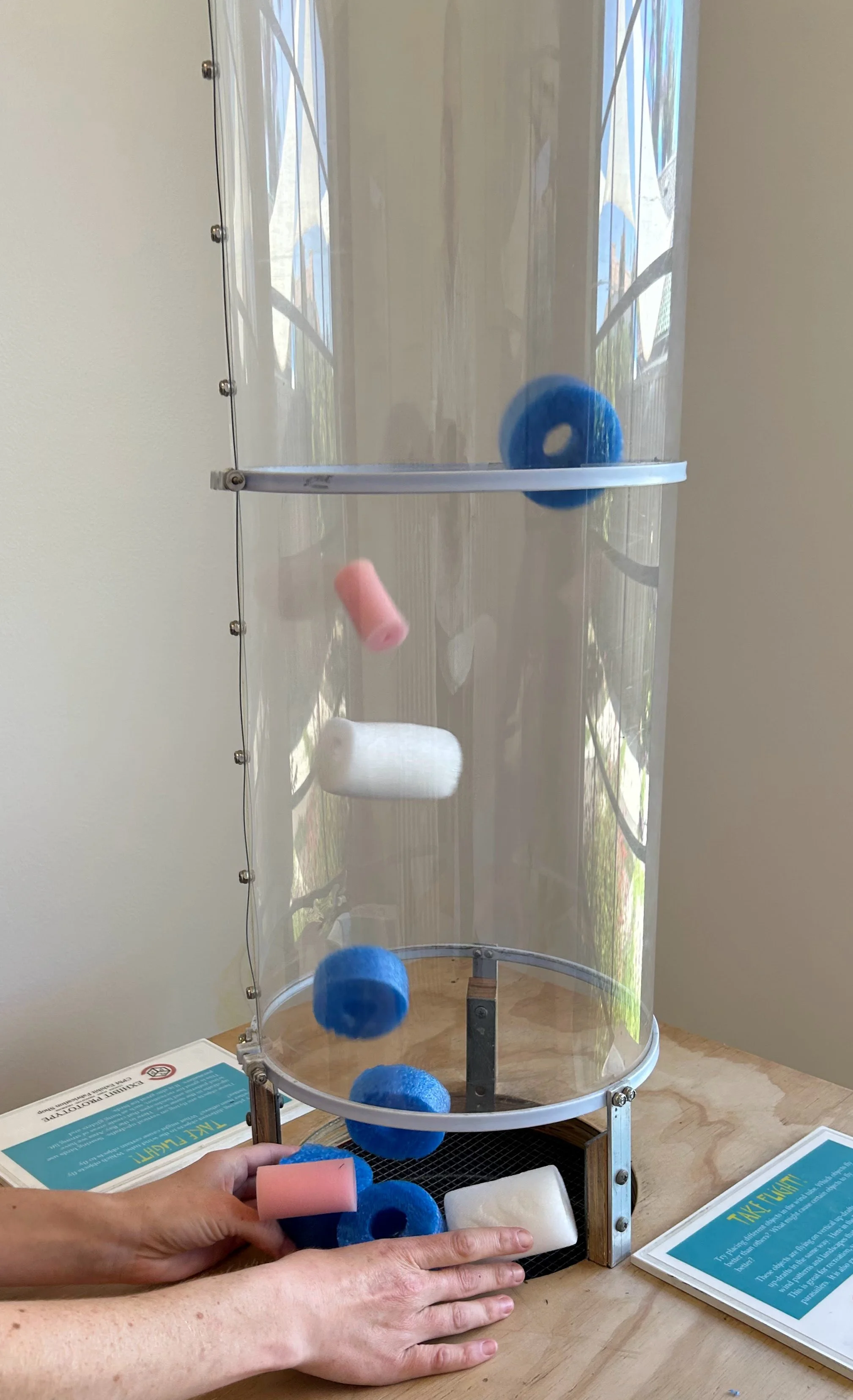 Personnel using a vertical cylindrical toy with foam and felt balls to demonstrate force and motion.