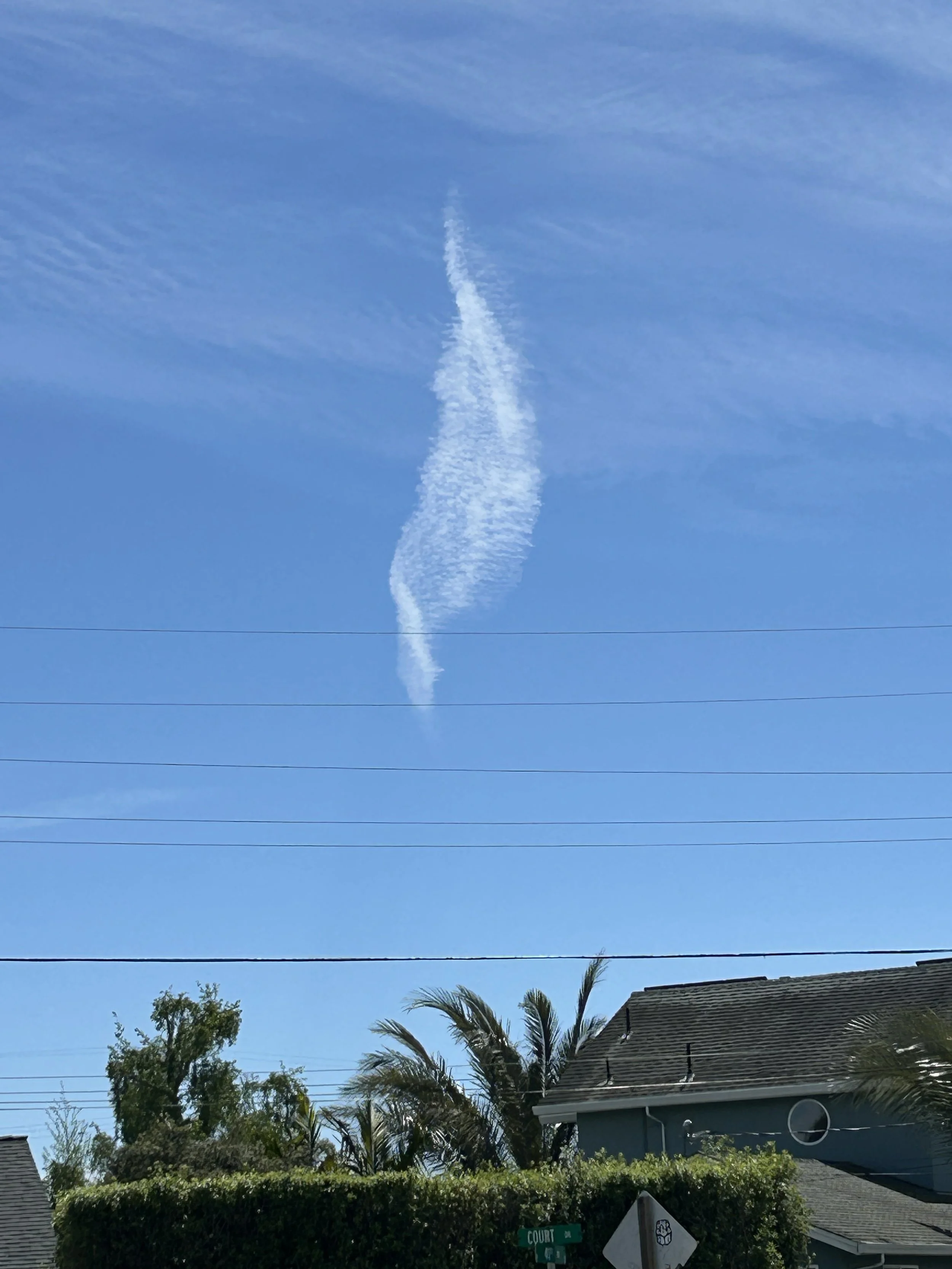 Blue sky with a contrail curling vertically above houses and trees.