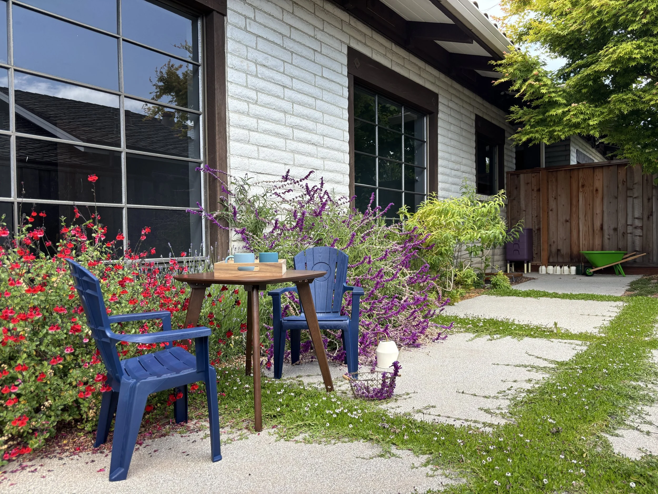 A cozy outdoor patio with two blue chairs around a small wooden table, surrounded by vibrant flowering plants and greenery, next to a house with large windows and a wooden fence.